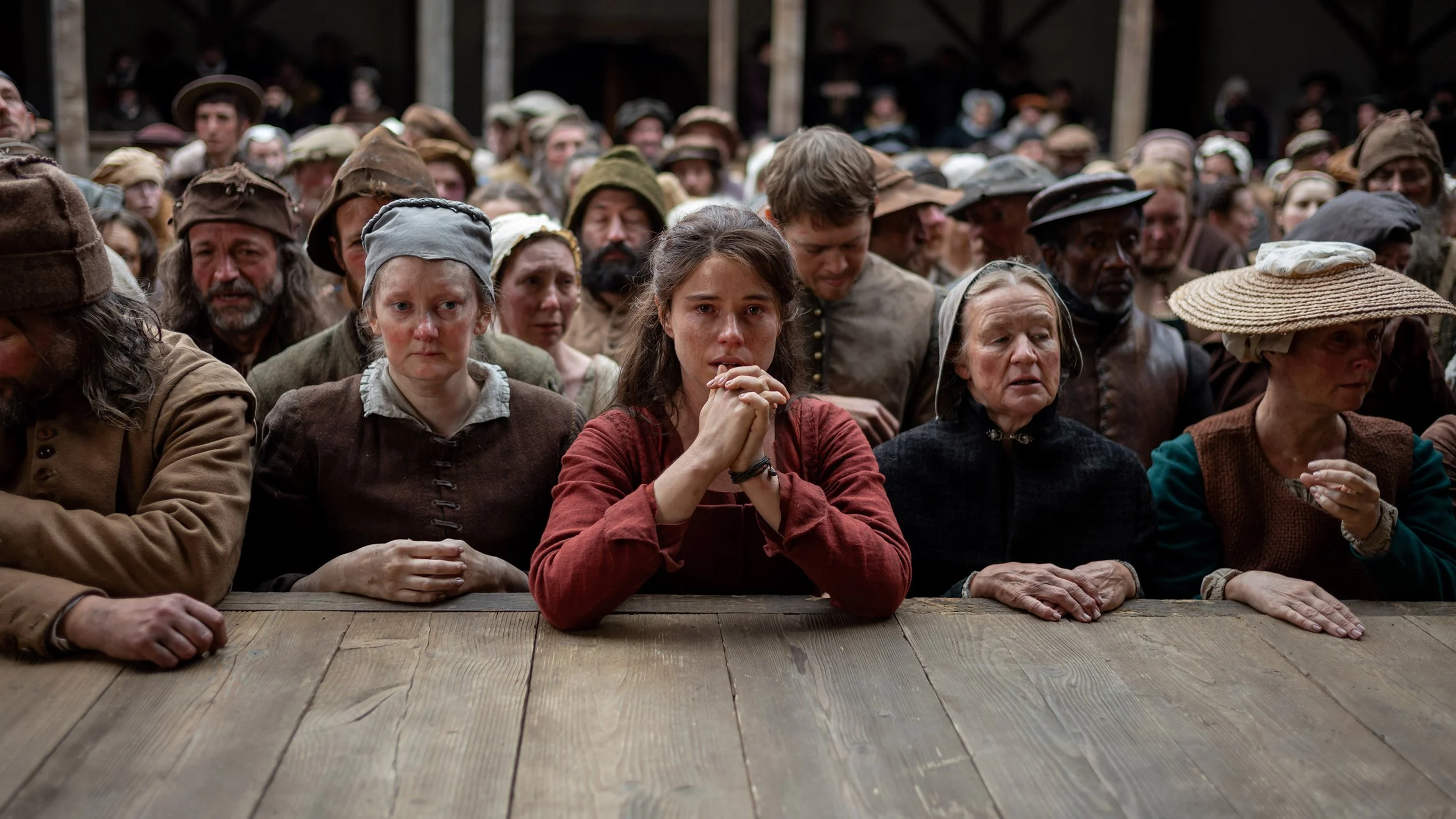 A crowd of people in period clothing stands closely packed at a wooden railing, with a woman at the center clasping her hands in grief while others around her watch solemnly, conveying shared sorrow and emotional intensity.