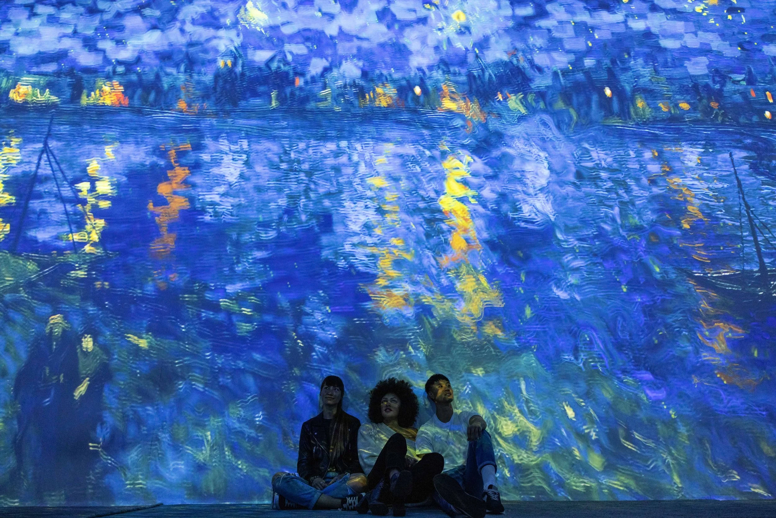 Three people sit together and look up at a Van Gough painting of sailboats in a harbor.