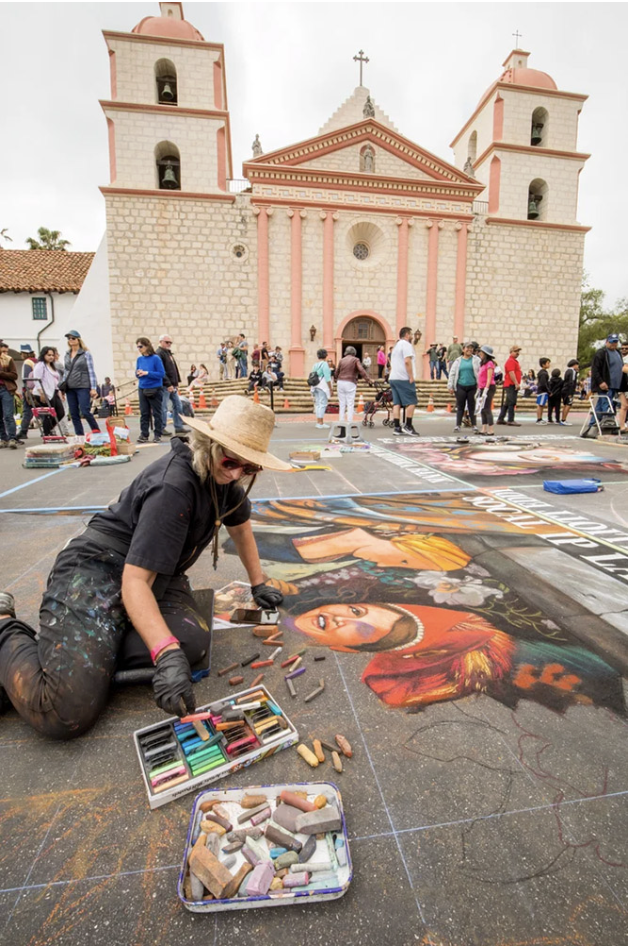 Person sitting next to chalk artwork on pavement at the I Madonnari Festival.