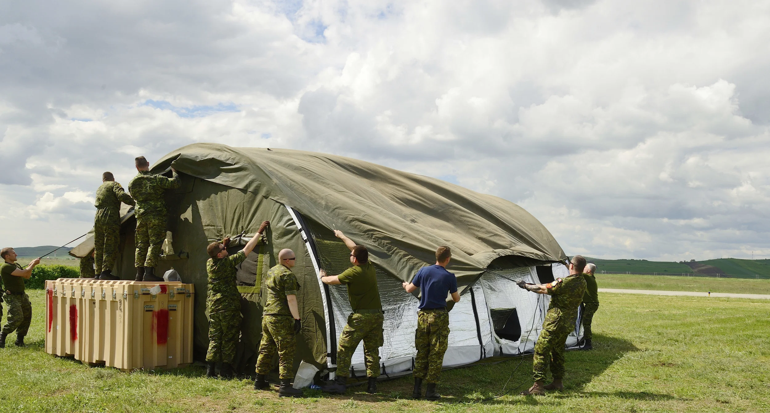 Members of Canadian Air Task Force put up tents in Câmpia Turzii, Romania, during NATO reassurance measures on May 10, 2014. The old-style canvas tents require a significant number of people and time to set up. New technology permits companies like HDT Global to develop easy-to-assemble, lightweight, durable deployable shelters that are easier and faster to set up yet can withstand harsh environments and be unsupplied for longer periods of time. (DND)