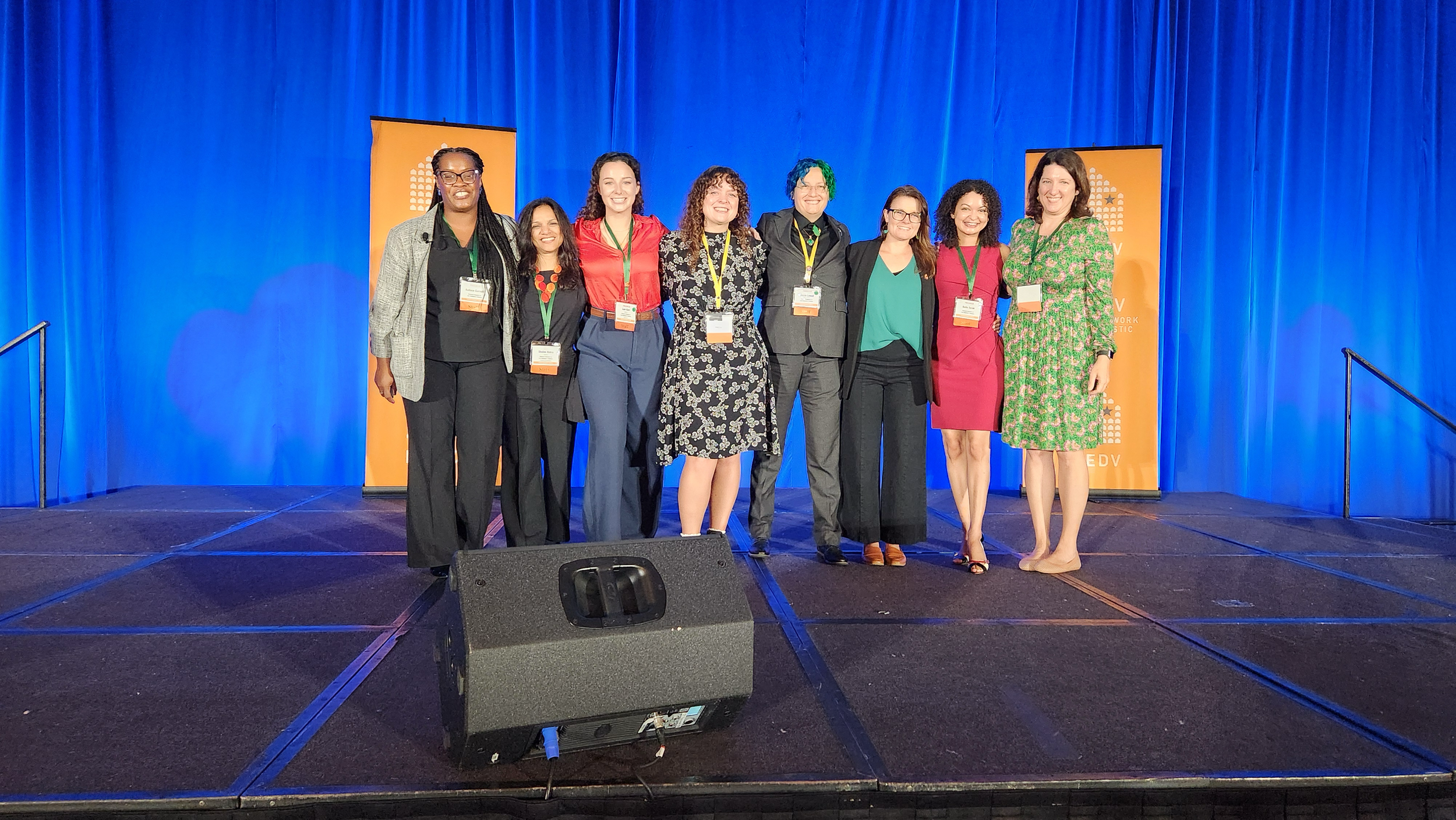 A photo of people dressed nicely and smiling on a stage - a tall Black woman, a short South Asian woman, a tall white woman, a medium-height white woman, a tall nonbinary person, a short white woman, a short Black woman, and a tall white woman
