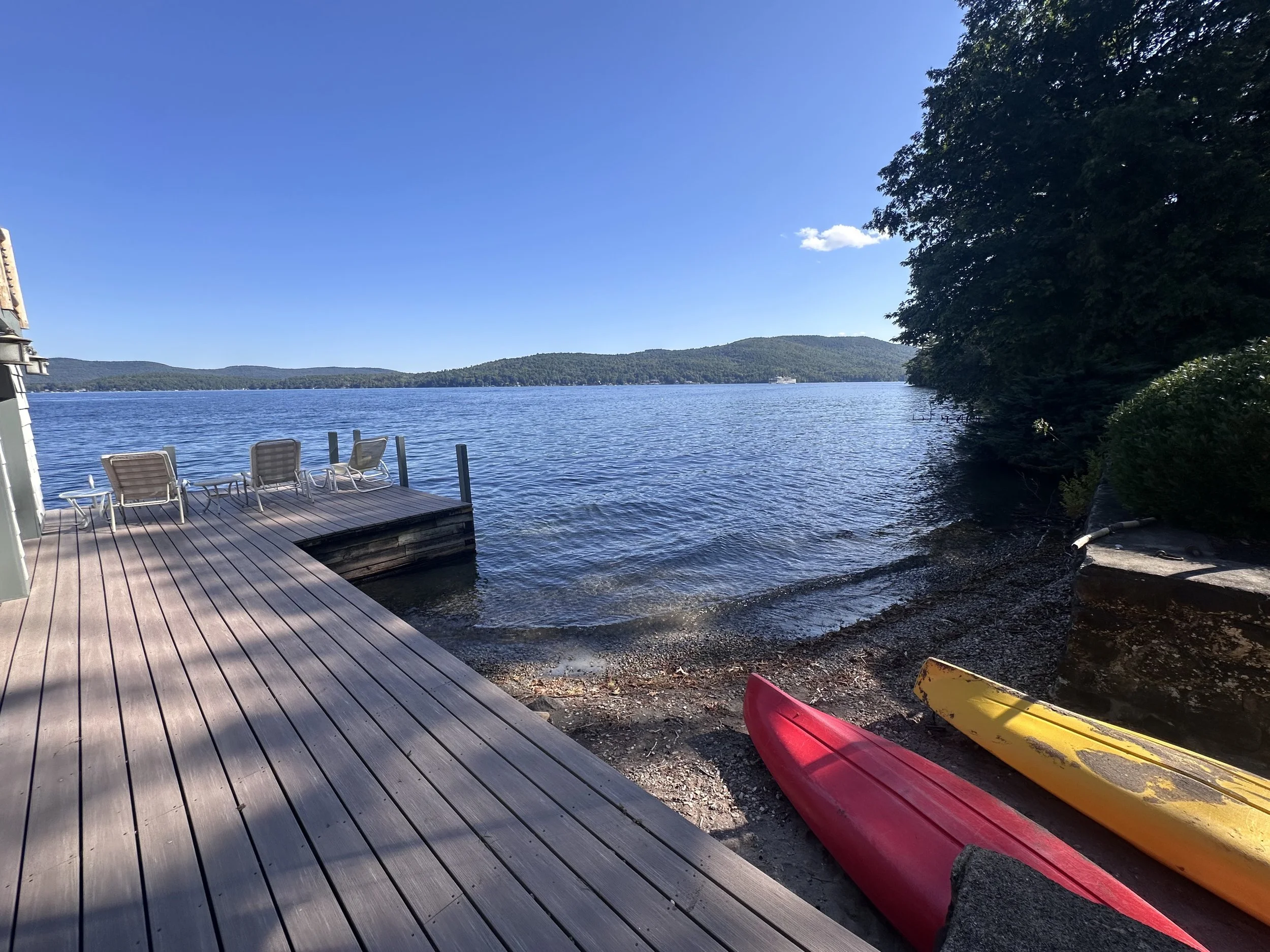 A lakeside scene with a wooden dock and several lounge chairs facing the water. Two kayaks, one red and one yellow, rest on the shore near the dock. The water is calm, surrounded by trees and distant hills under a clear blue sky.