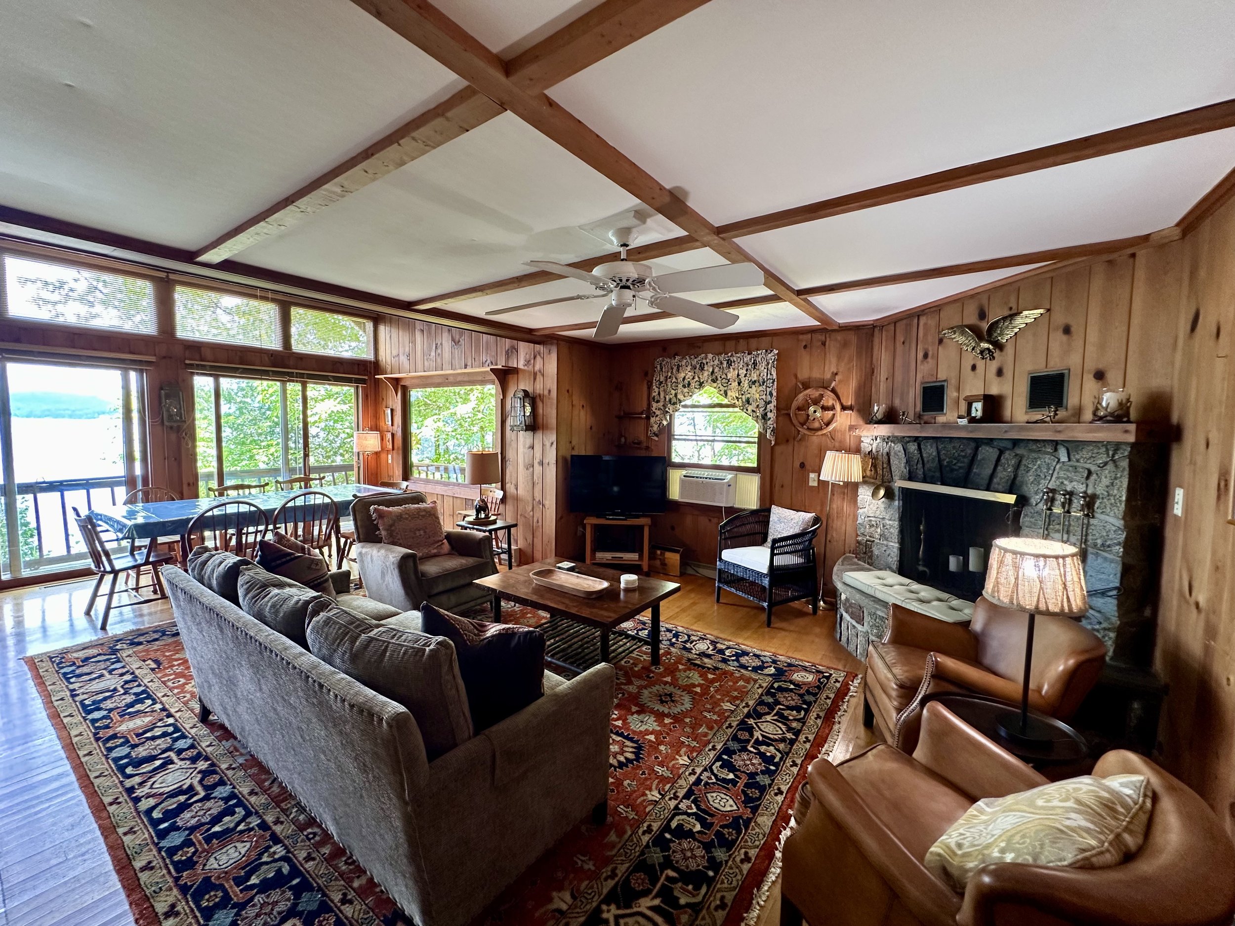 Living room with wood paneled walls, large windows overlooking nature, a fireplace, and various chairs and a sofa, decorated with a patterned rug and vintage decor.