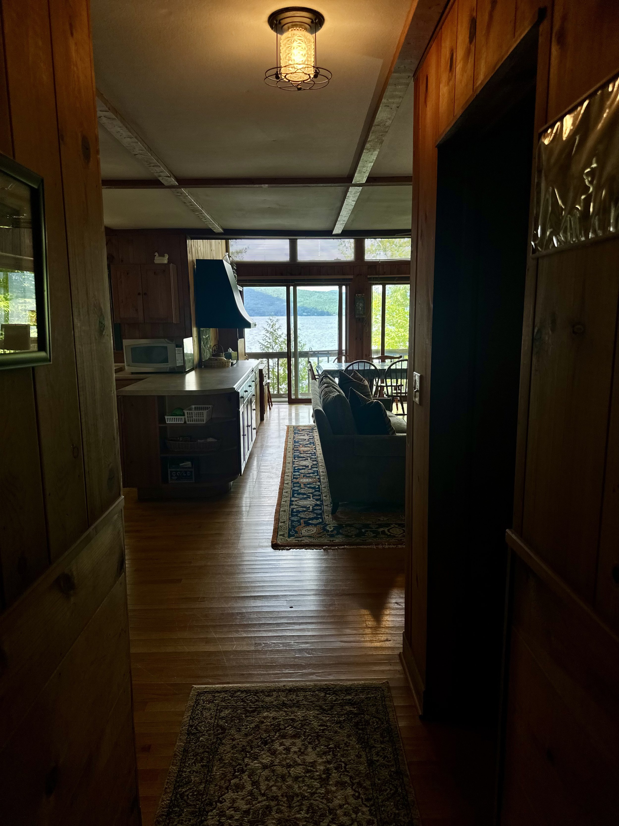 View from a wooden hallway into a living room with large windows showing a lake and trees outside, a sofa, a patterned rug, and a dining table.