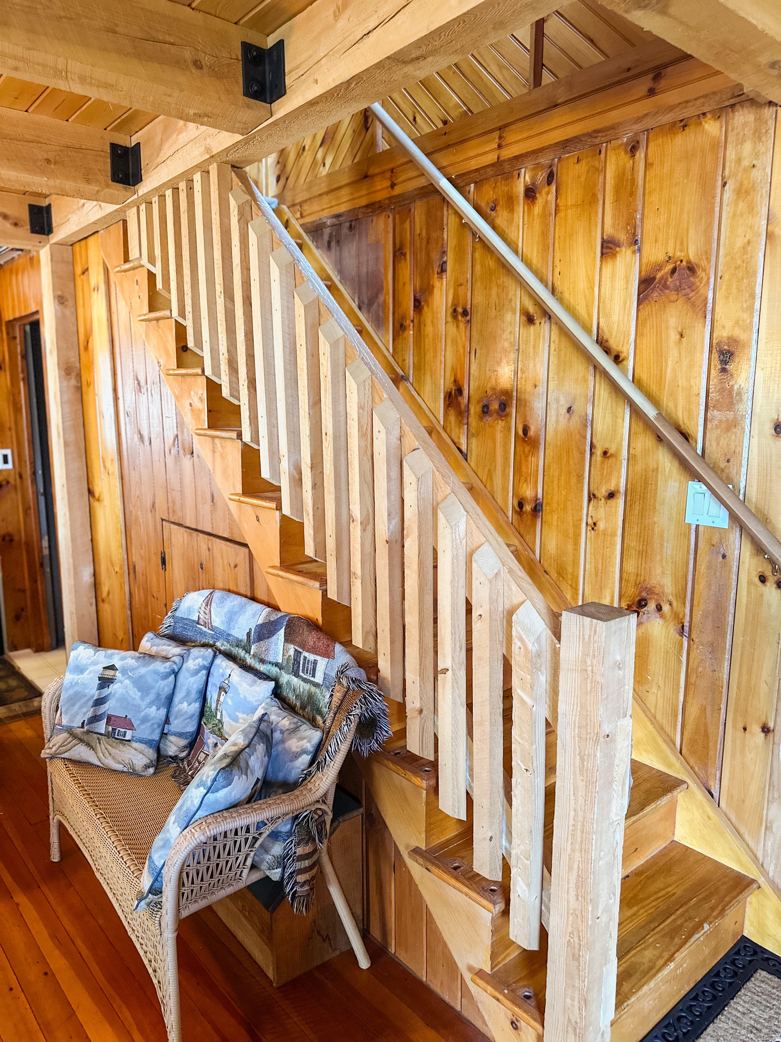 The image shows a wooden staircase in a cozy room with wooden paneling on the walls. A wicker chair with a lighthouse-themed blanket is placed in front of the staircase.