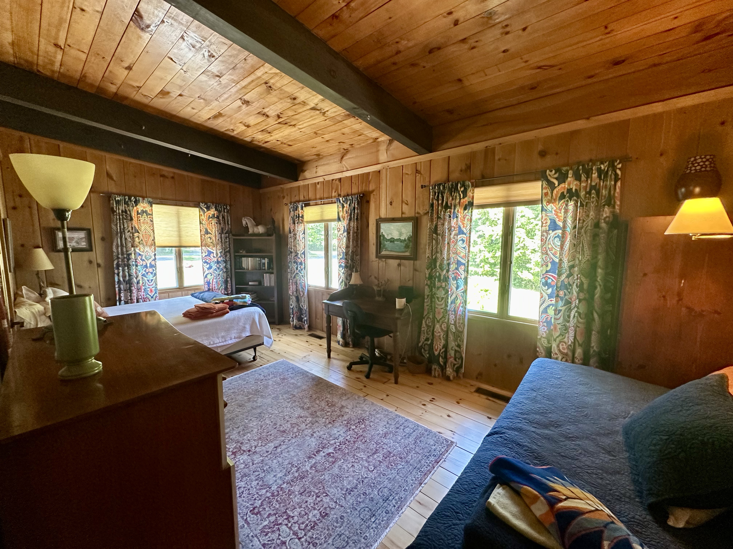 A cozy wooden-paneled bedroom with two windows, a bed with sentient objects, a dark blue couch, a desk with a chair, and colorful patterned curtains.