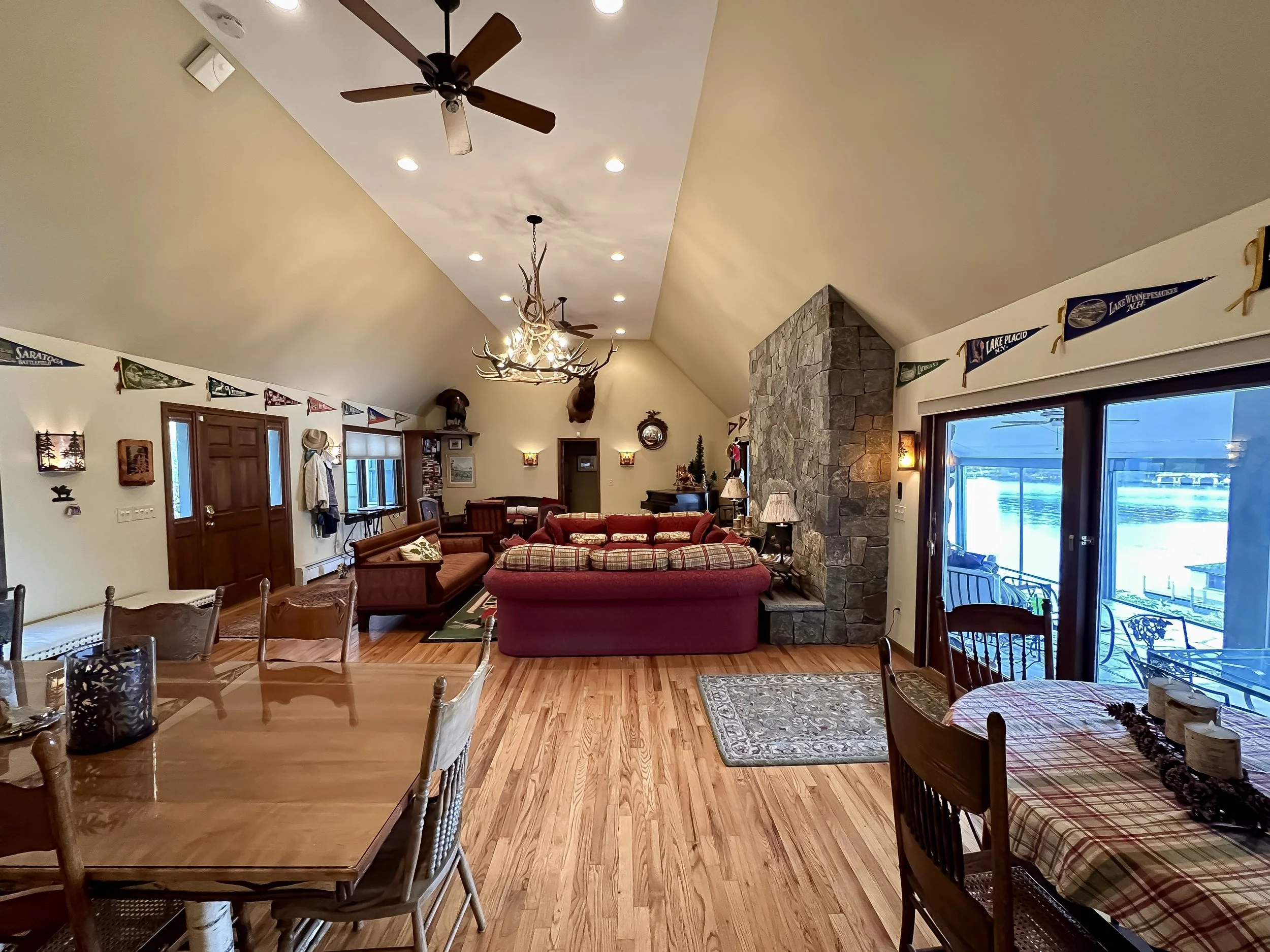 Living room with vaulted ceiling, ceiling fans, and hardwood flooring. Contains a stone fireplace, multiple sofas, a wine rack, flag banners, and a sliding glass door leading to a porch with outdoor furniture.