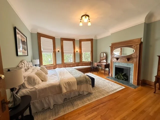 Bedroom with large bed, multiple pillows, nightstand with lamp, window with natural light, a fireplace with mirror, a chair, and hardwood floors.