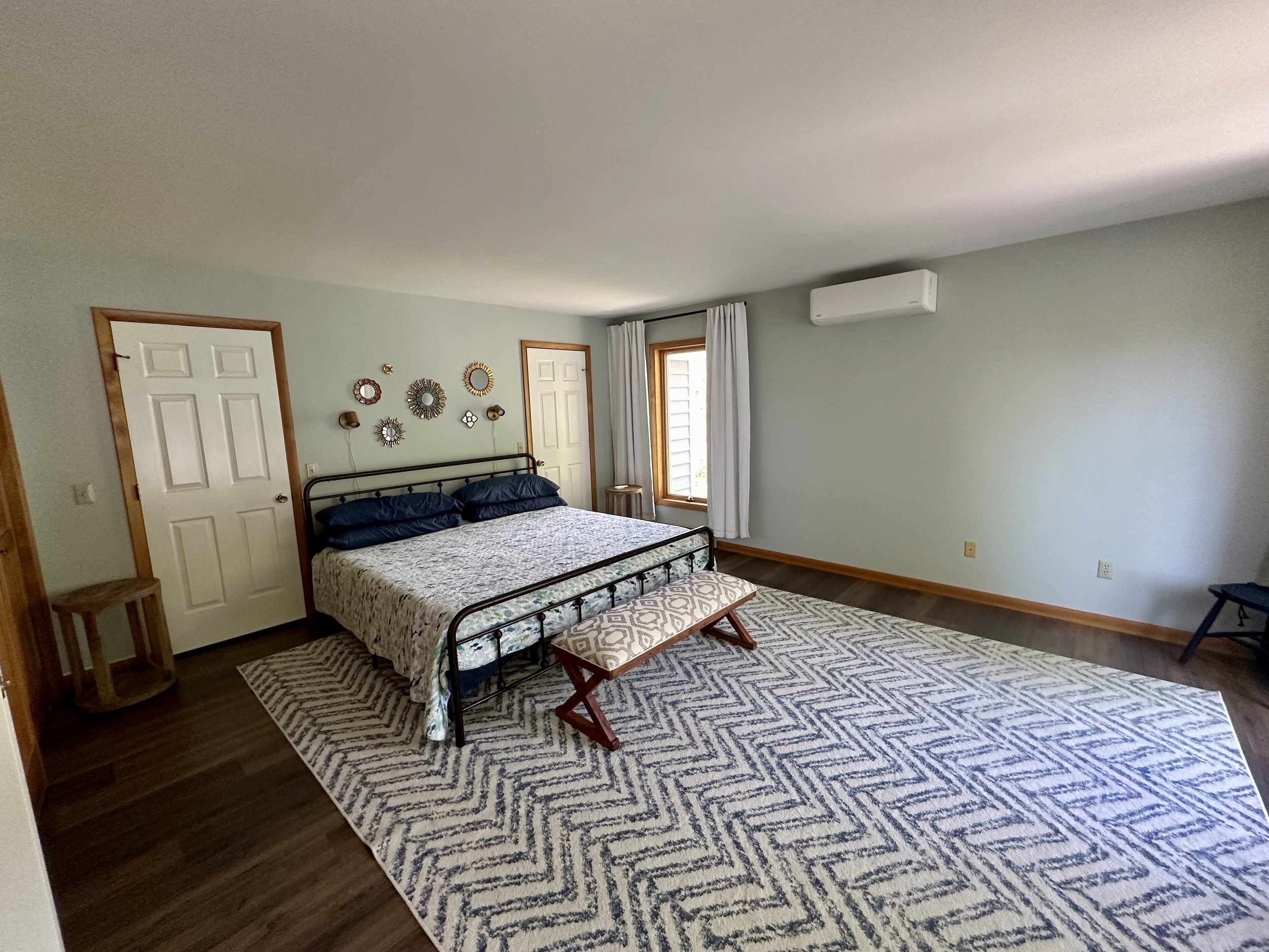 Bedroom with a black metal bed, navy blue pillows, patterned bedspread, wooden bench at foot of bed, two doors, a window with white curtains, wall decor, air conditioning unit, wooden floors, and a large beige and navy patterned area rug.
