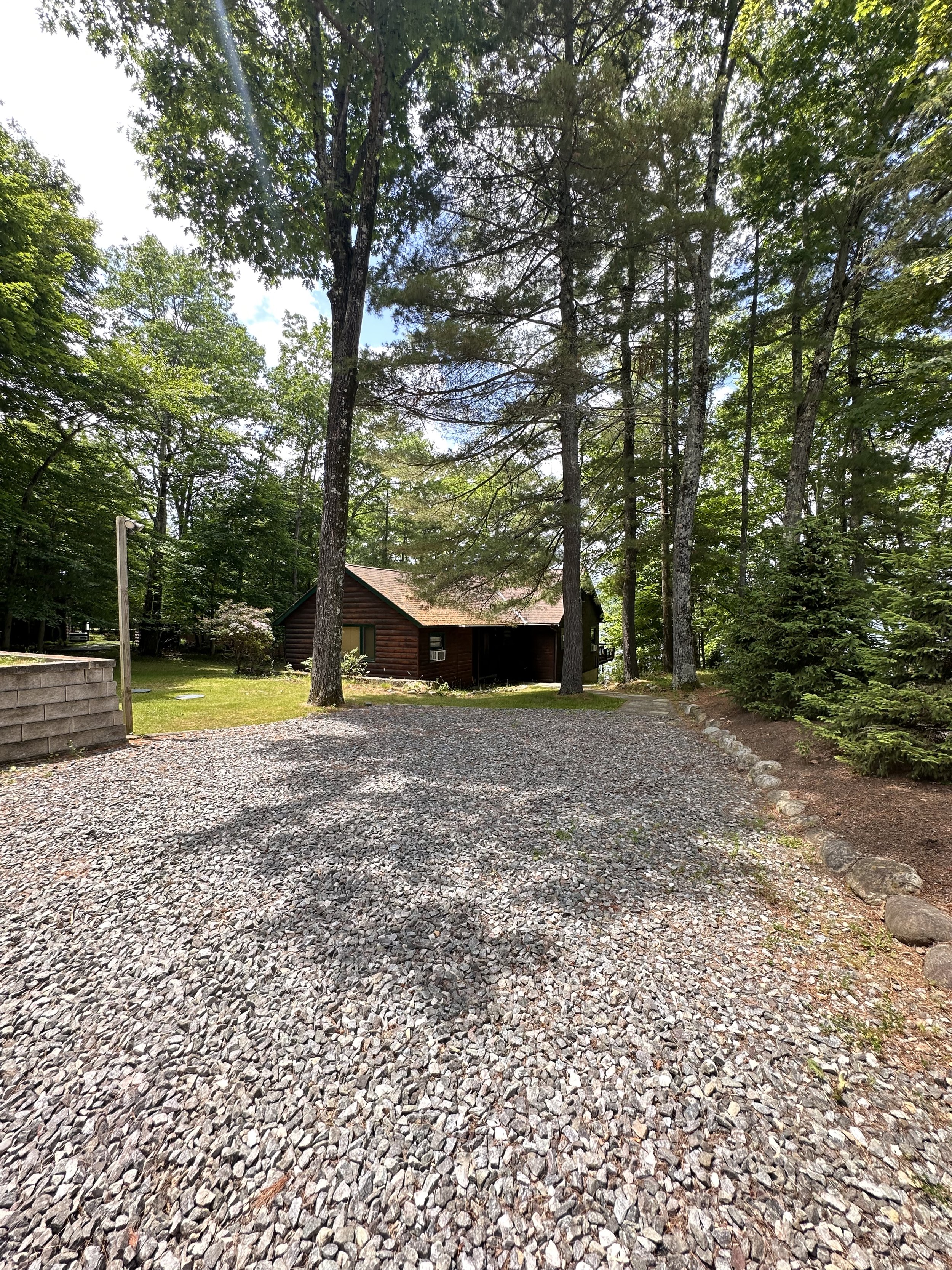 A gravel driveway leading to a small wooden house among trees on a clear day with blue sky.