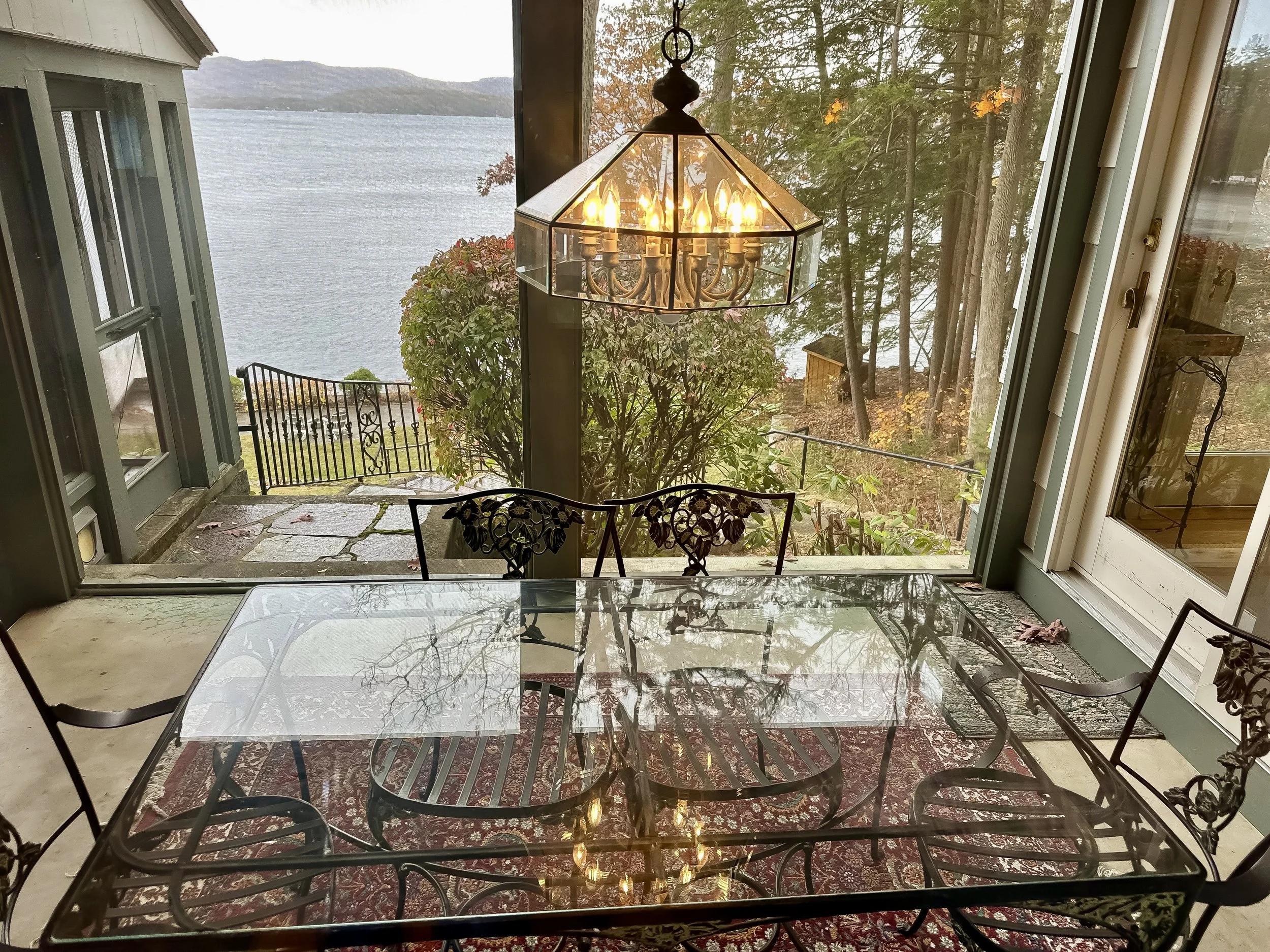 A glass-top dining table with wrought iron chairs inside a sunroom, overlooking a lakeside view with mountains and trees. A decorative chandelier hangs above the table. Outside, there is a railing, a bush, and a deck with a stone pathway leading to a