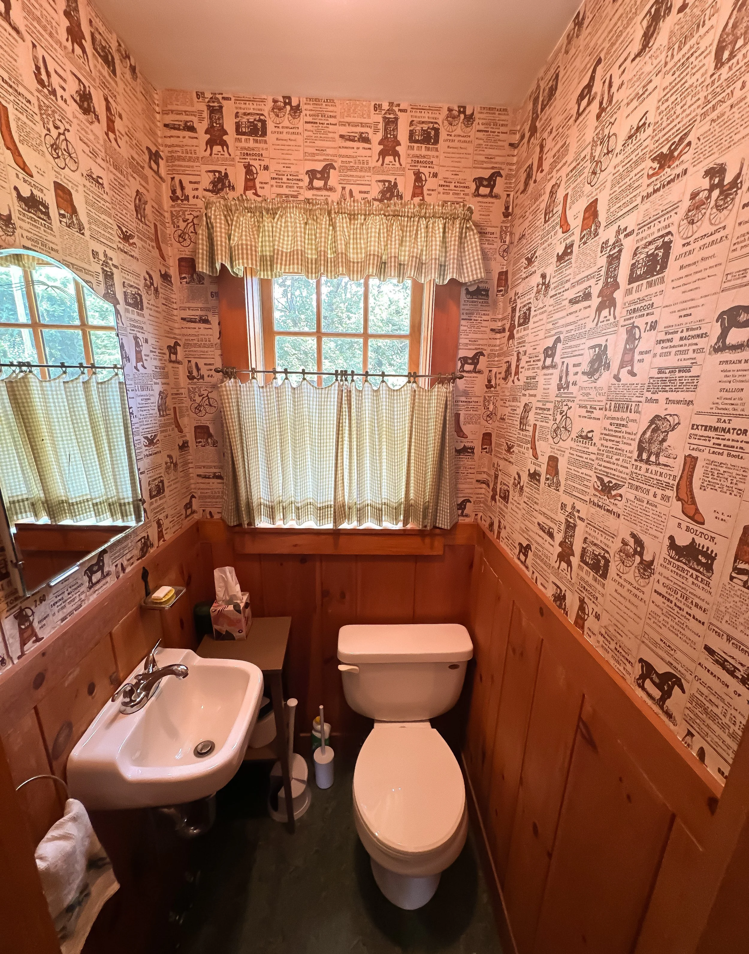 Small rustic bathroom with wooden wainscoting, wallpaper featuring vintage advertisements and illustrations, a small sink, a toilet, a mirror, a small side table with tissue box, and green checkered curtains on two windows.
