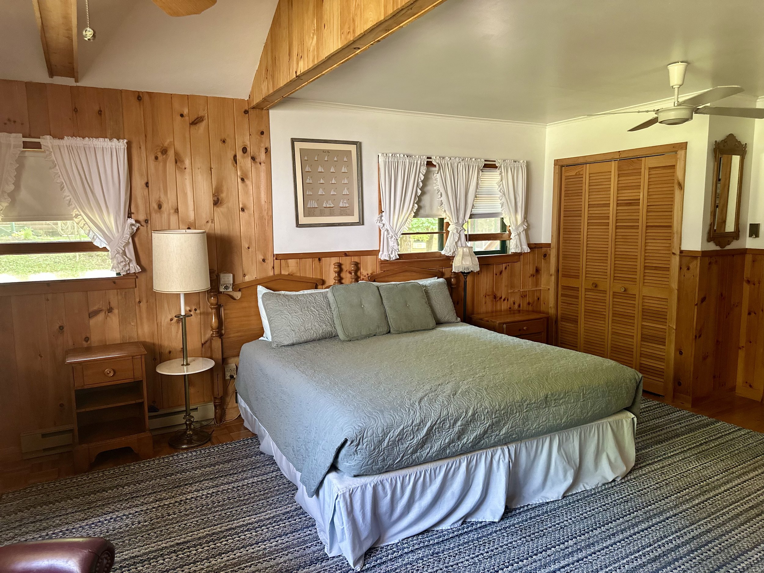A cozy bedroom with wood-paneled walls, a large bed with green and white bedding, wooden nightstands, a lamp, a framed picture of sailboats on the wall, multiple windows with white curtains, a built-in wooden closet, a ceiling fan, and a colorful are