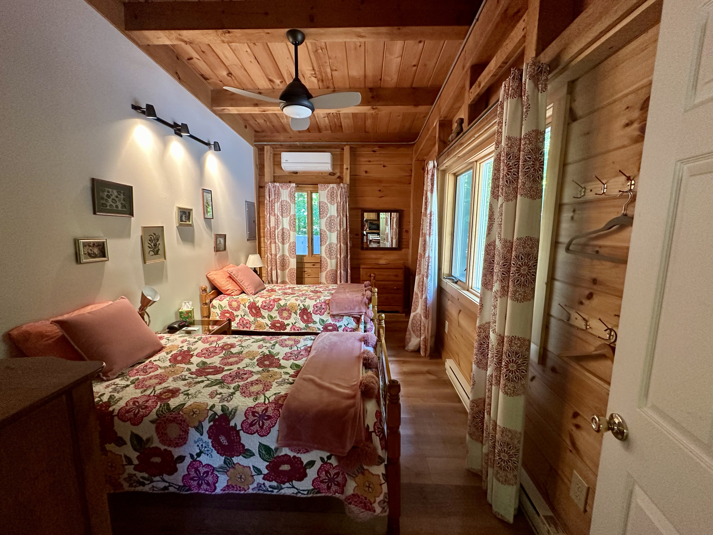 A cozy bedroom with two twin beds featuring floral bedspreads and pink pillows, wooden walls, a ceiling fan, and windows with patterned curtains.