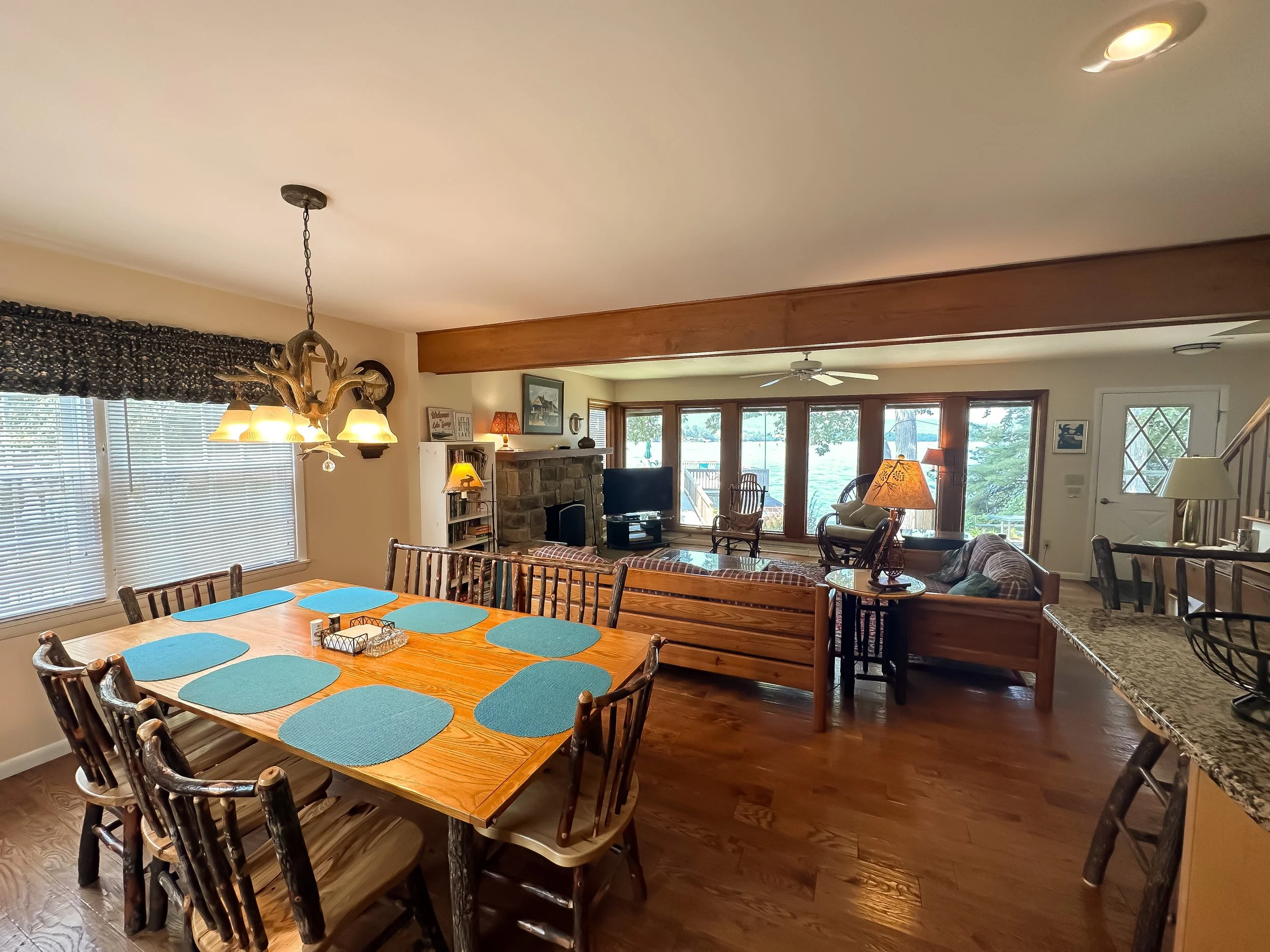 Open-concept dining and living room area with wooden furniture, a stone fireplace, large windows overlooking water, and cozy seating