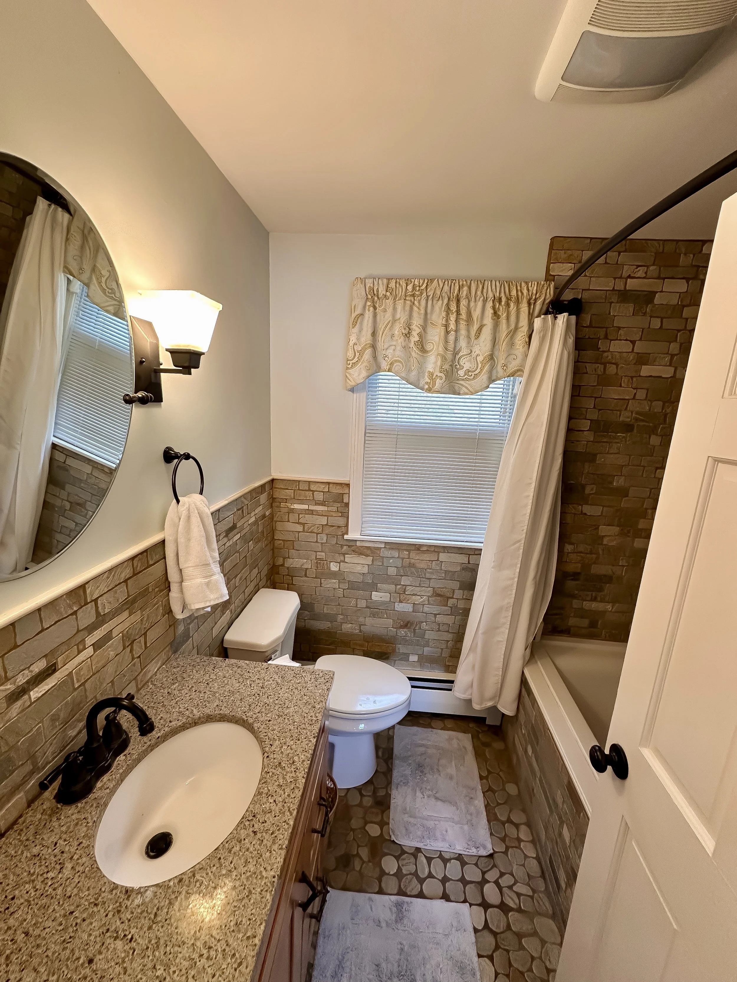 Bathroom with a granite countertop sink, oval mirror, wall sconce, towel ring with towel, toilet, small window with blinds and valance, shower curtain, brick wall accents, pebble-style tile flooring, and bath mats.
