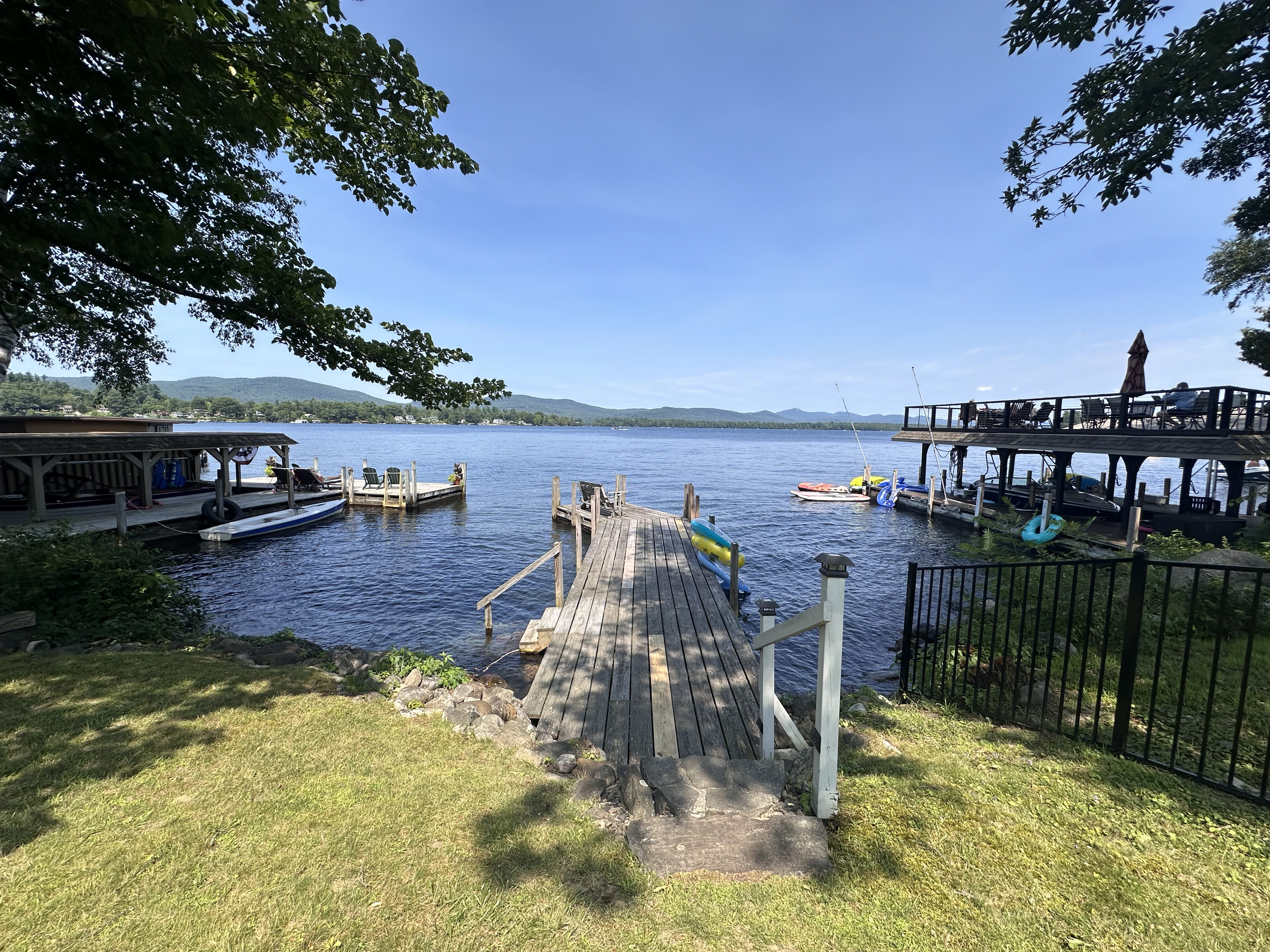 View of a lake with a wooden dock extending into the water, surrounded by boats, kayaks, and a lakeside deck, under a clear blue sky with mountains in the distance.