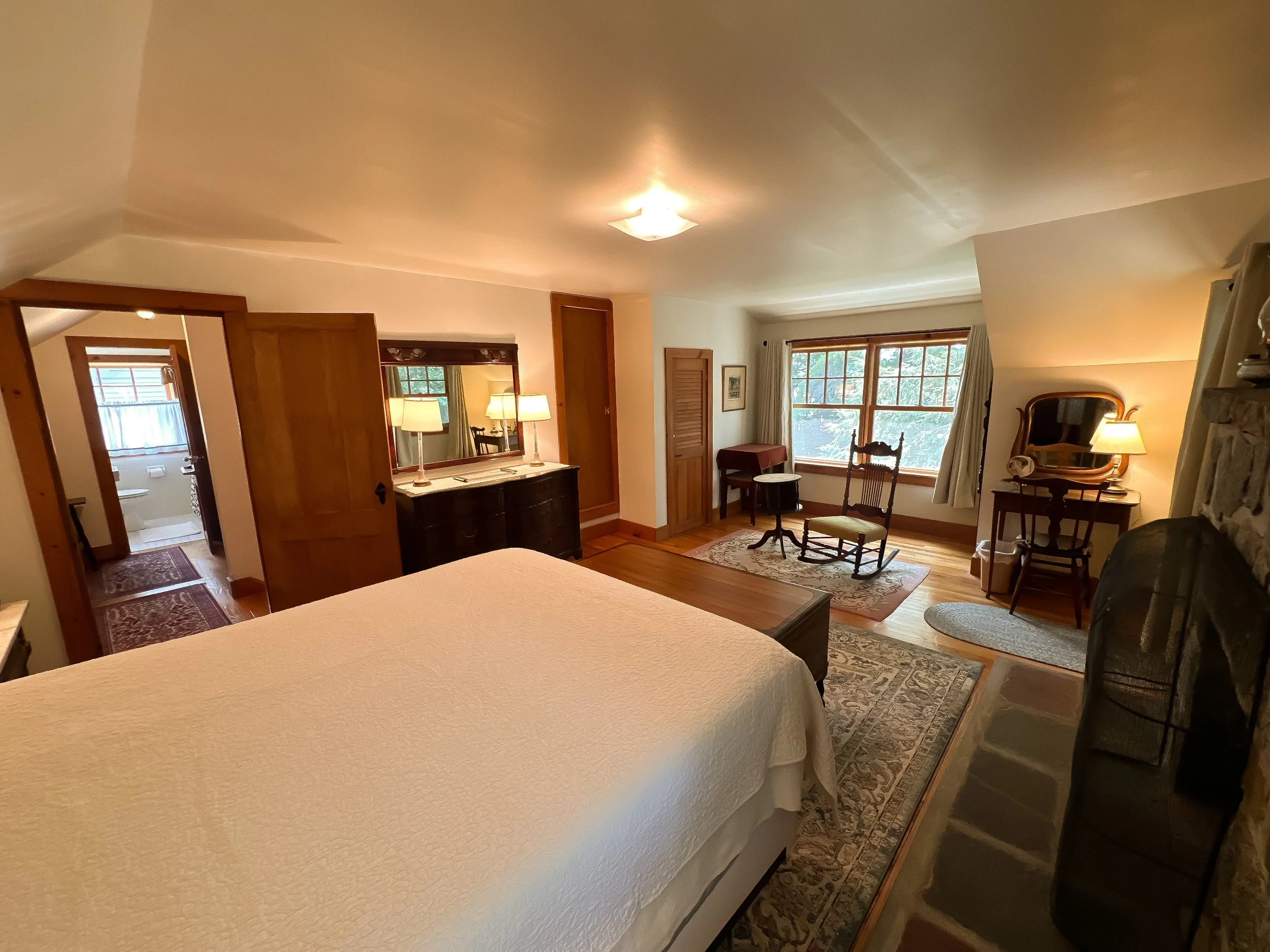 Cozy bedroom with a large bed, wooden furniture, a rocking chair, a dresser with lamps, a window with curtains, and a view into a bathroom with a toilet and window.