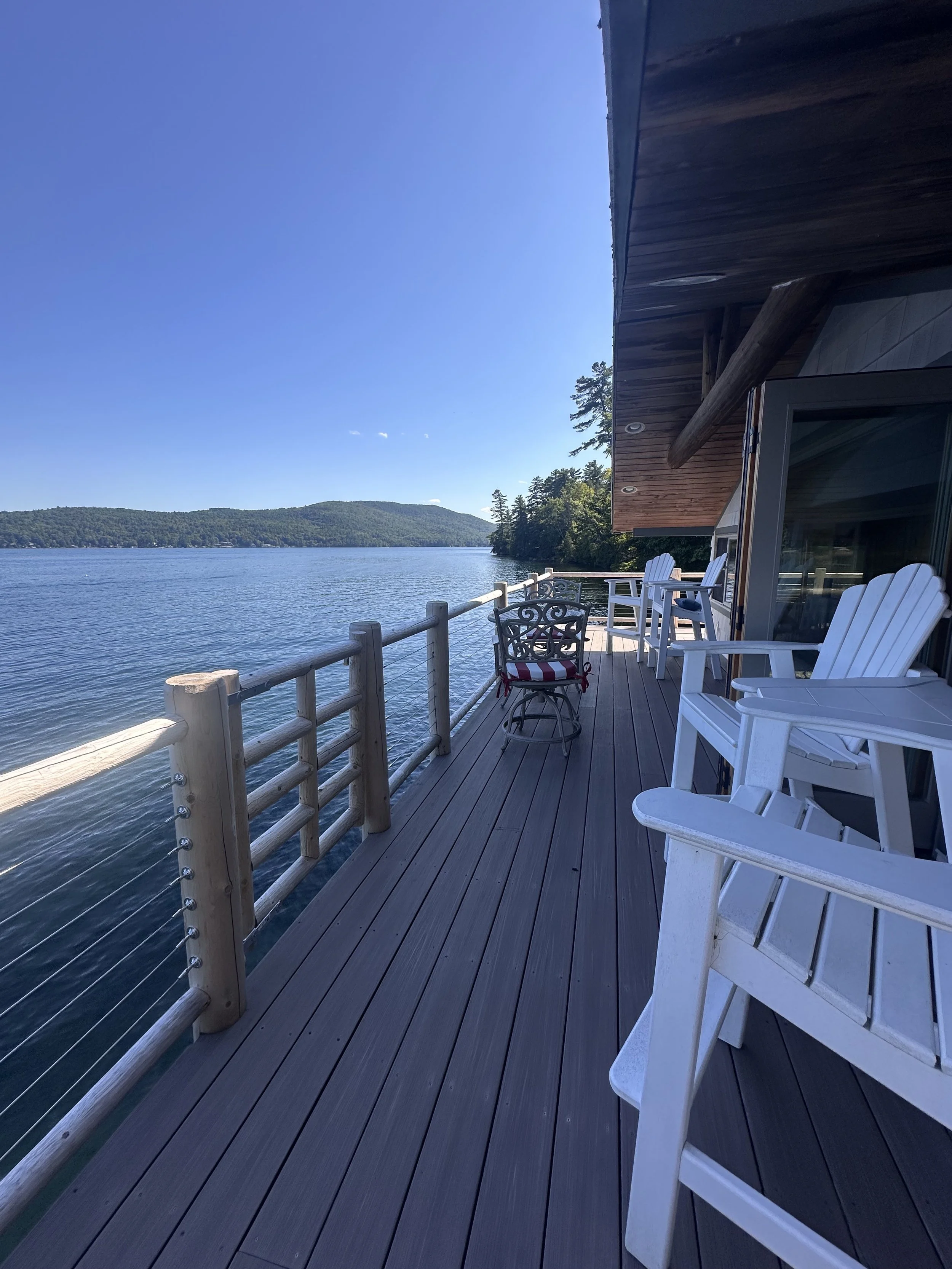 A spacious wooden deck overlooking a large lake with lush green hills in the background. The deck has white Adirondack chairs and a small decorative metal chair with a red, white, and blue cushion. Clear blue sky and calm water create a peaceful outd
