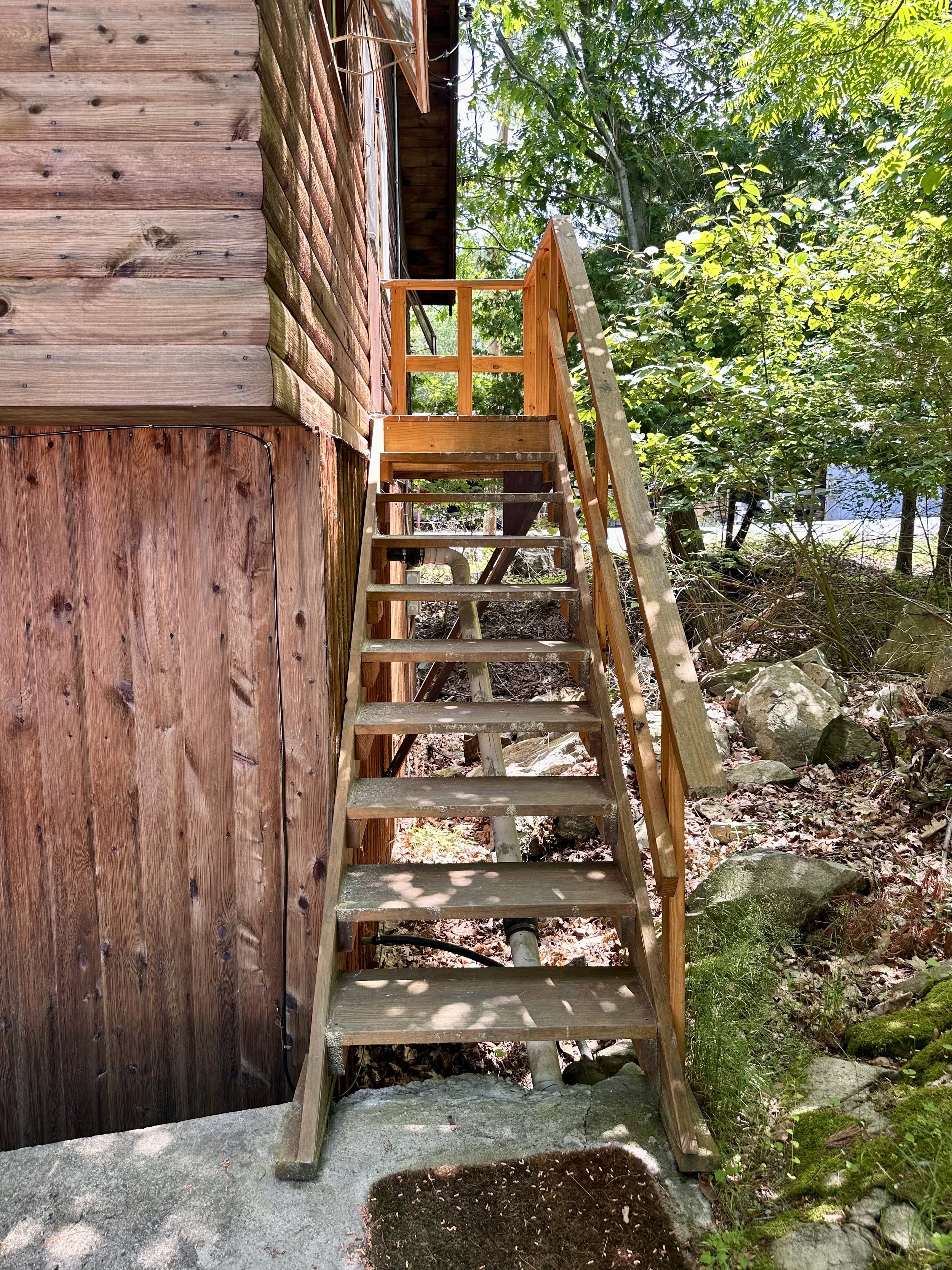 Wooden staircase leading up to a deck attached to a wooden house, surrounded by trees and rocks.