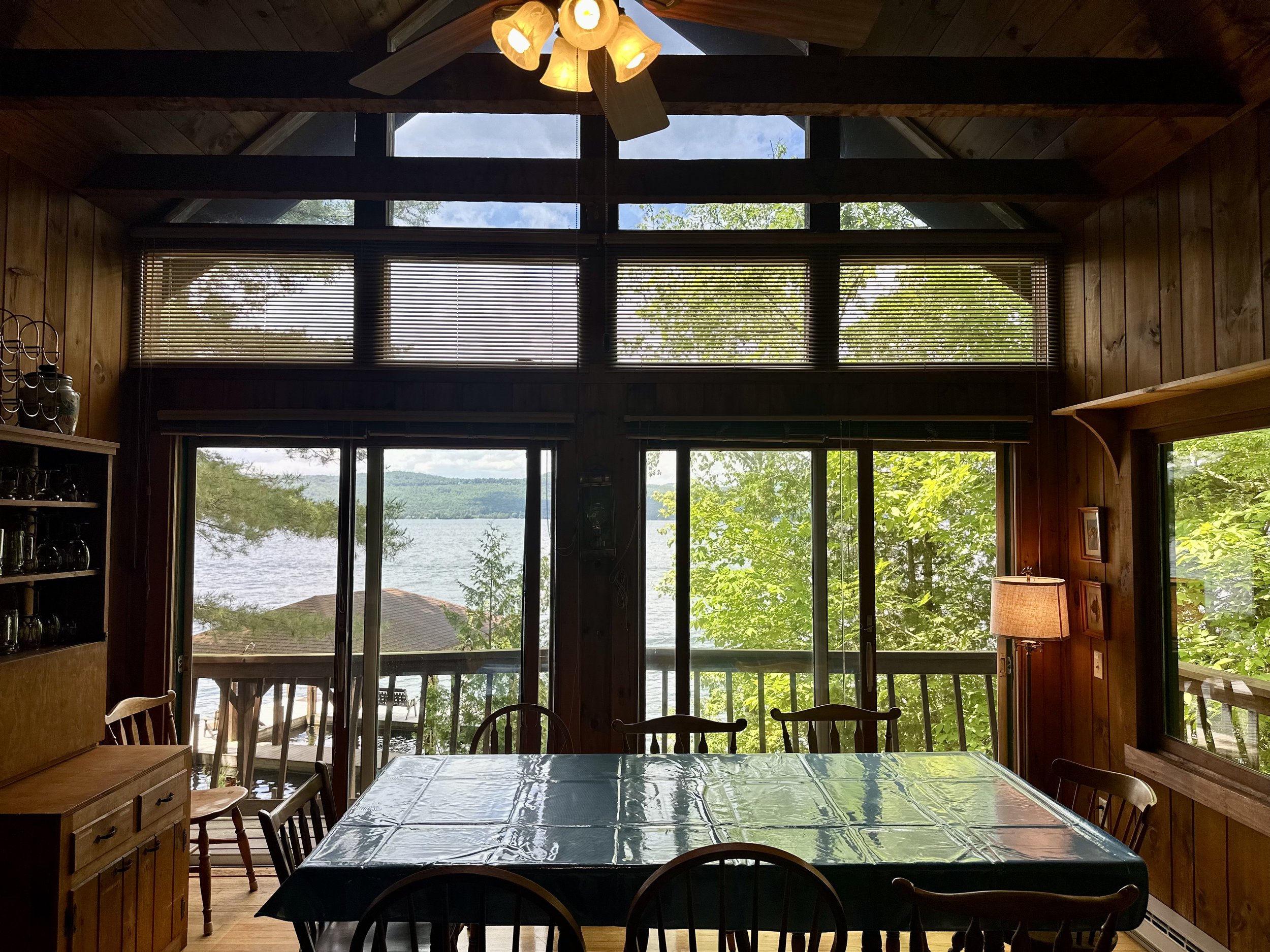 Interior of a wooden dining room with large windows showing a lake and trees outside, a table with a plastic tablecloth, and a sliding glass door leading to a balcony with outdoor furniture.