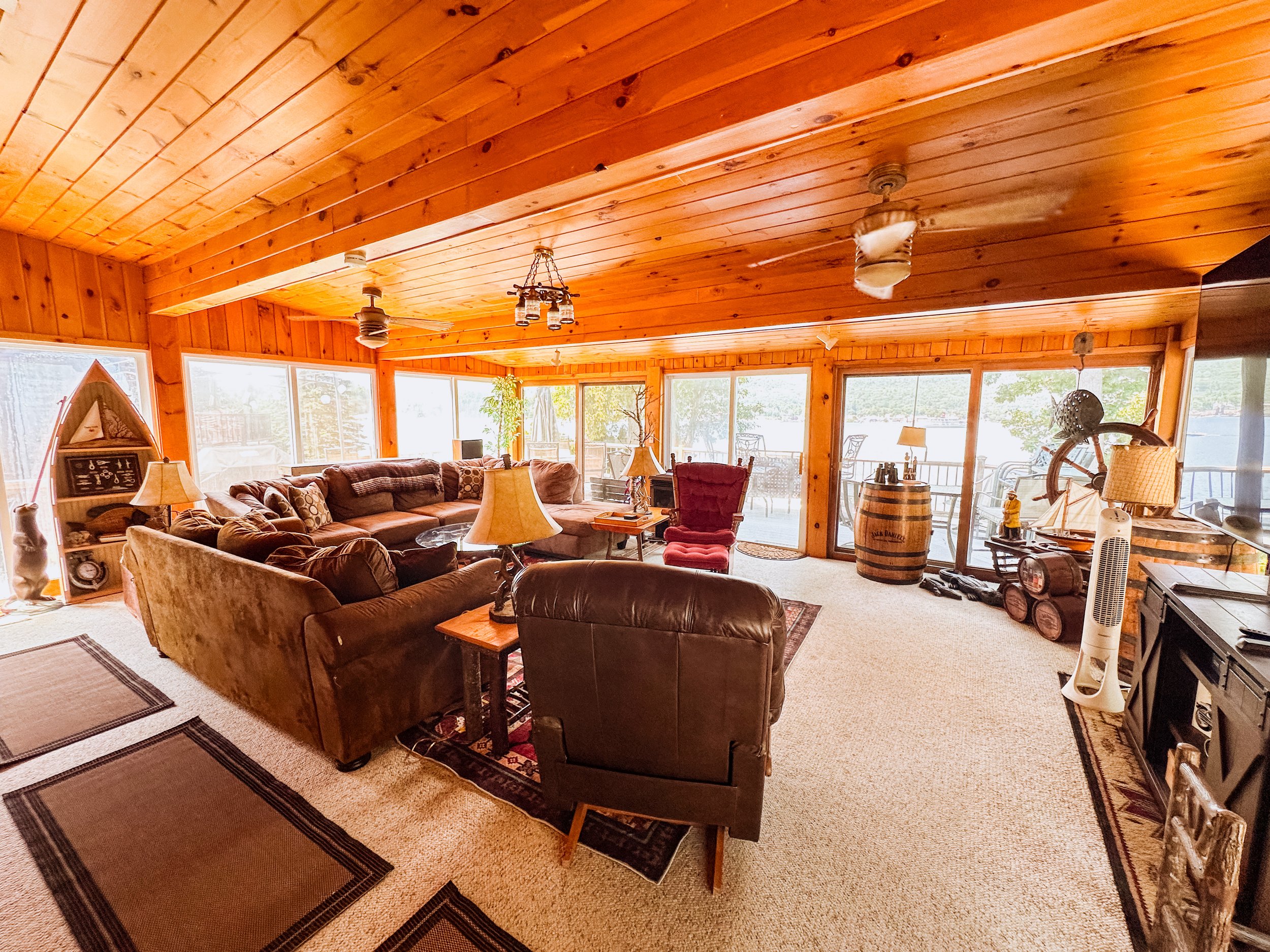 A cozy living room with wooden paneled ceiling and walls, featuring a large brown sectional sofa with decorative pillows, a red armchair, lamps, and various decorative items, with large windows and a balcony door letting in natural light.