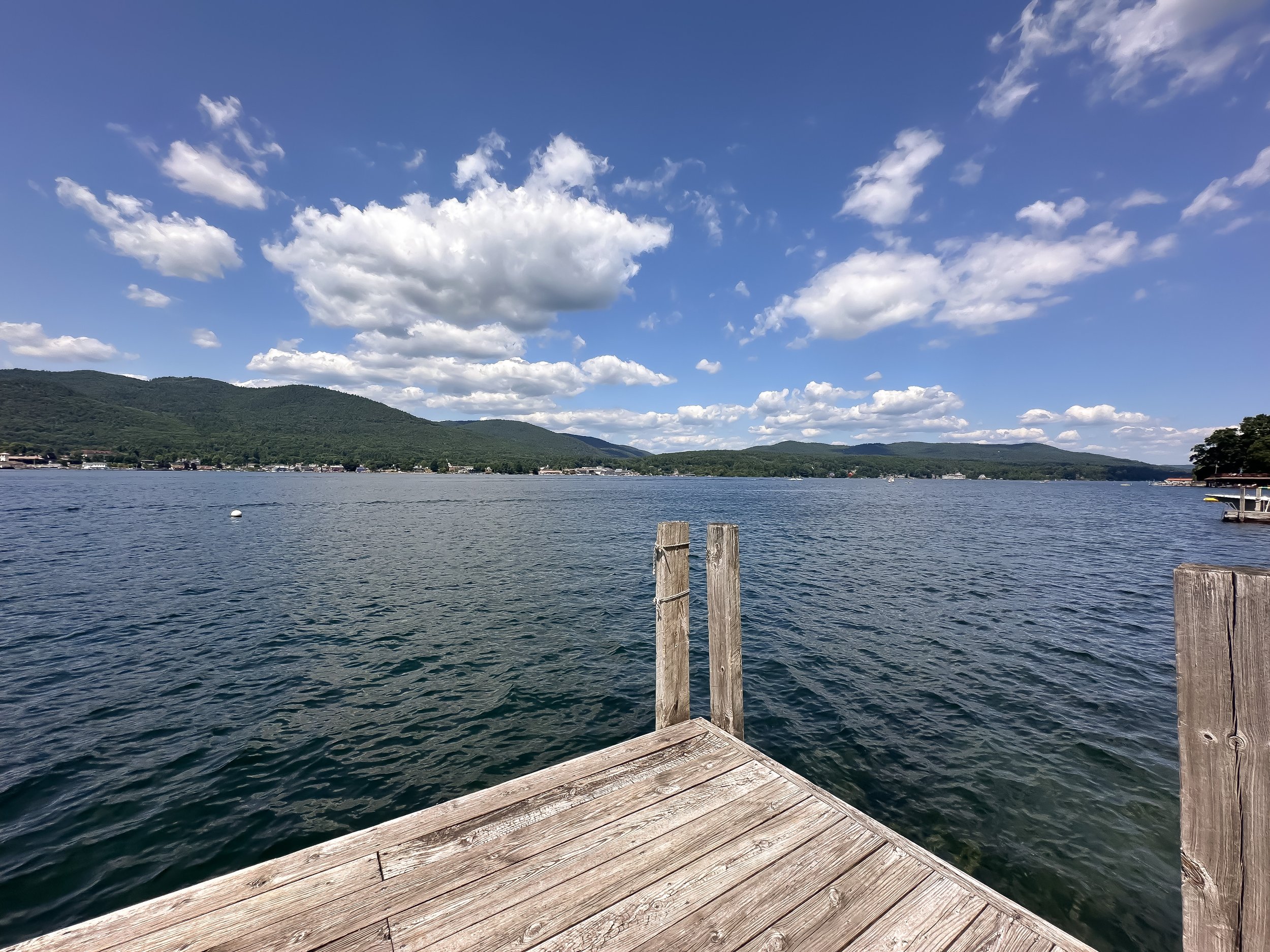 View of a lake with mountains and a partly cloudy blue sky in the background, seen from a wooden dock.
