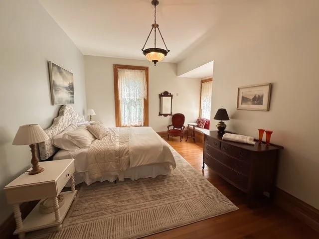 A cozy bedroom with a white bed, nightstand, and lamps on each side, a wooden dresser with decorative items, a window with lace curtains, and a sitting area with a red chair and paintings on the walls.