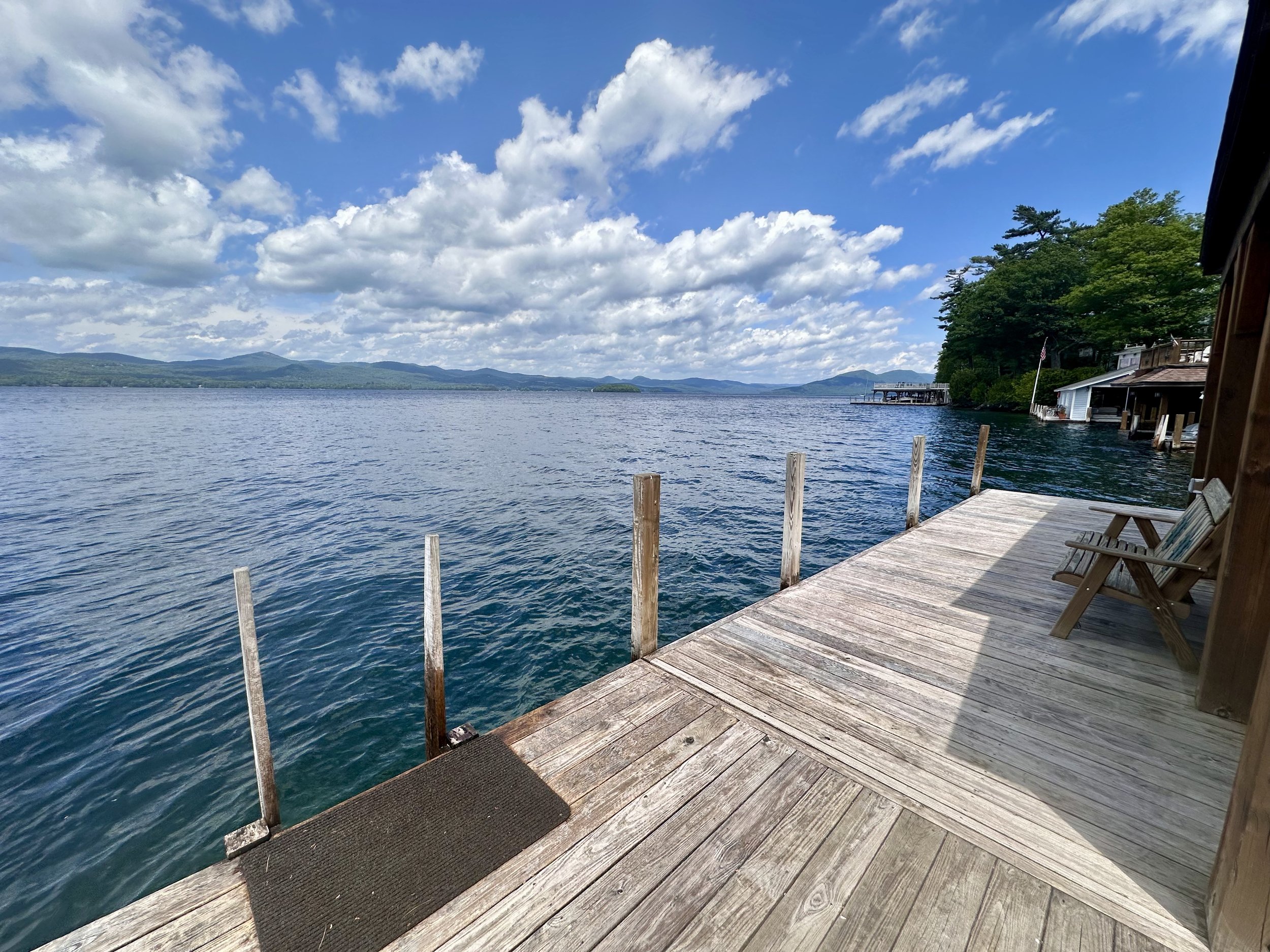 View of a wooden dock extending over a calm lake with mountains in the background and partly cloudy sky.