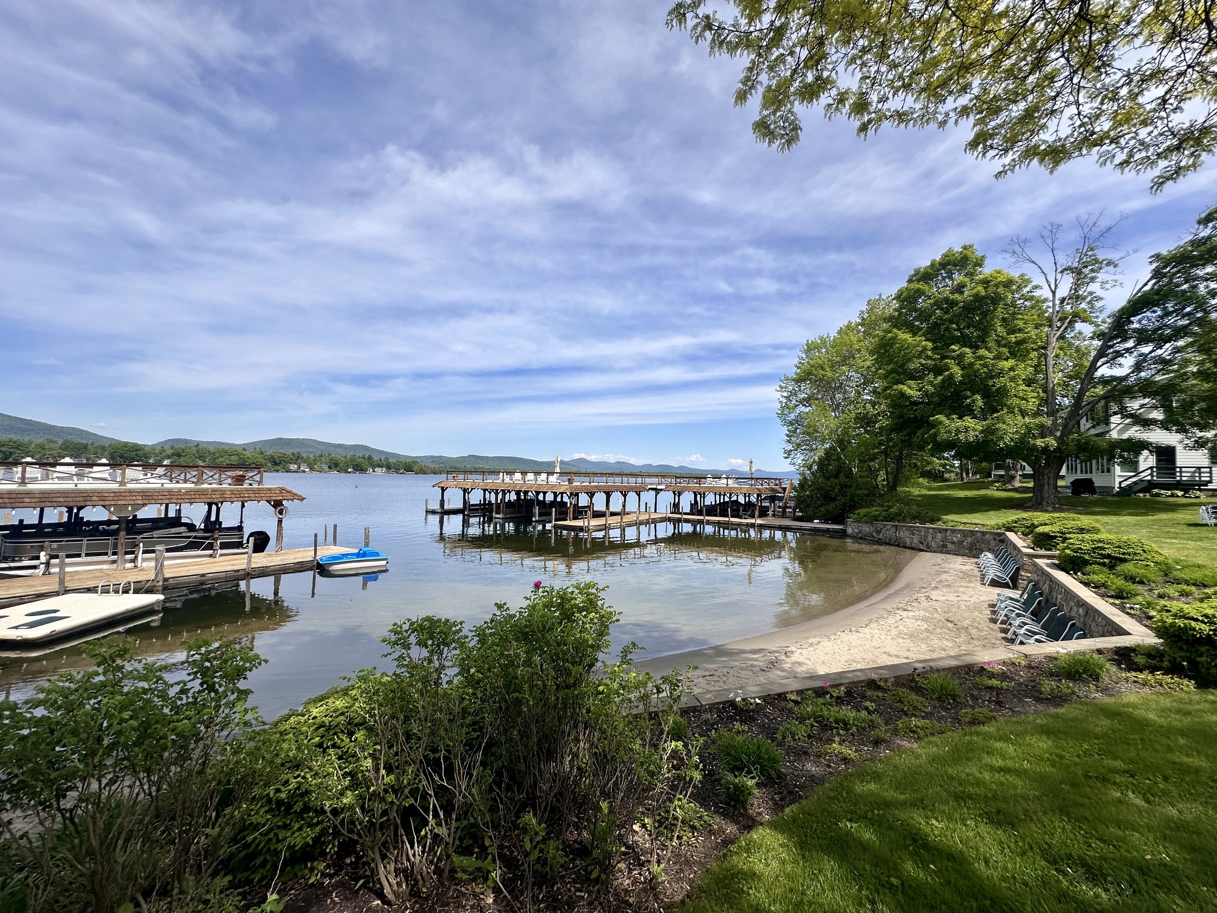 Scenic lakeside view with a sandy beach, green trees, a dock with boats, and a house in the background under a blue sky with some clouds.