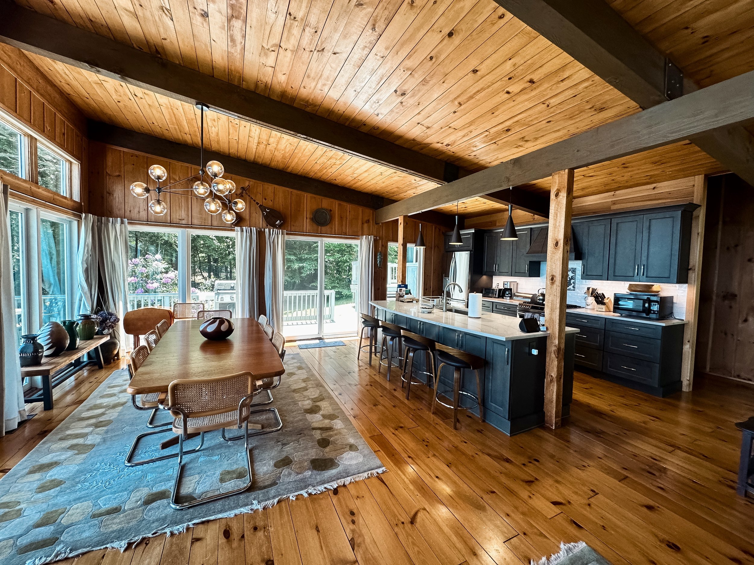 Open-concept kitchen and dining area in a rustic home with wooden walls, ceiling, and floors, featuring a large dining table with chairs, a kitchen island with barstools, and sliding glass doors leading outside.