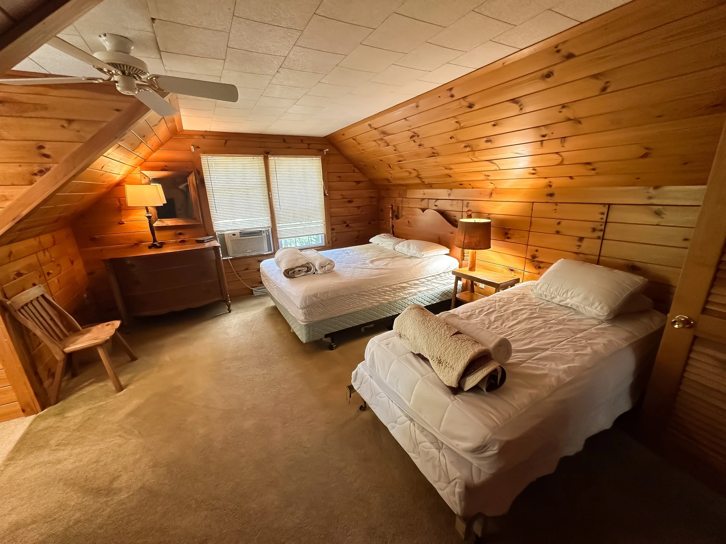 A bedroom with wooden paneling on the walls and ceiling, two beds with white sheets and pillows, a small wooden nightstand with a lamp, a wooden chair, and a window with blinds and an air conditioning unit.
