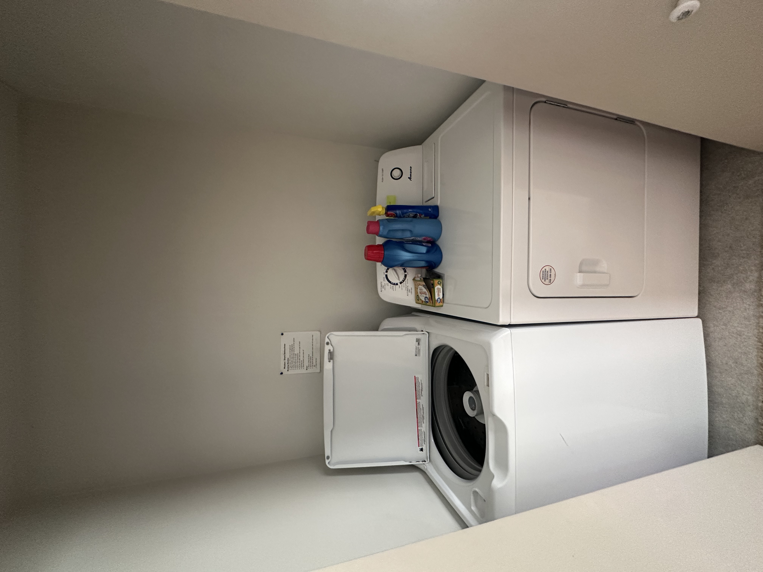 A laundry room with a stacked washer and dryer, with laundry detergent bottles and a box of laundry pods on top of the washer.