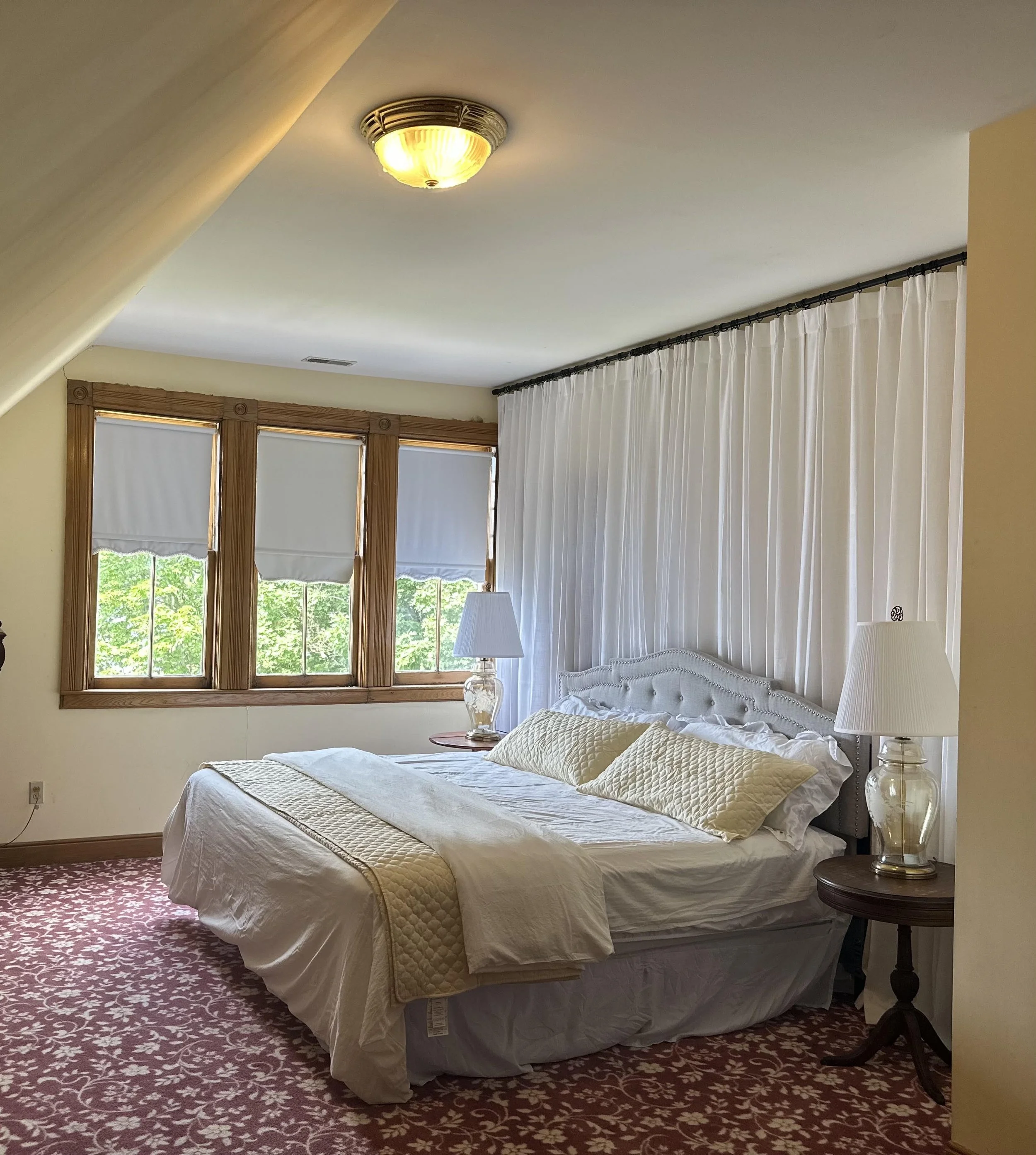 A cozy bedroom with a large bed covered in white and yellow bedding. There are two lamps on nightstands beside the bed, and a window with wooden framing and white shades. White curtains hang along the window, with a view of green trees outside. The c