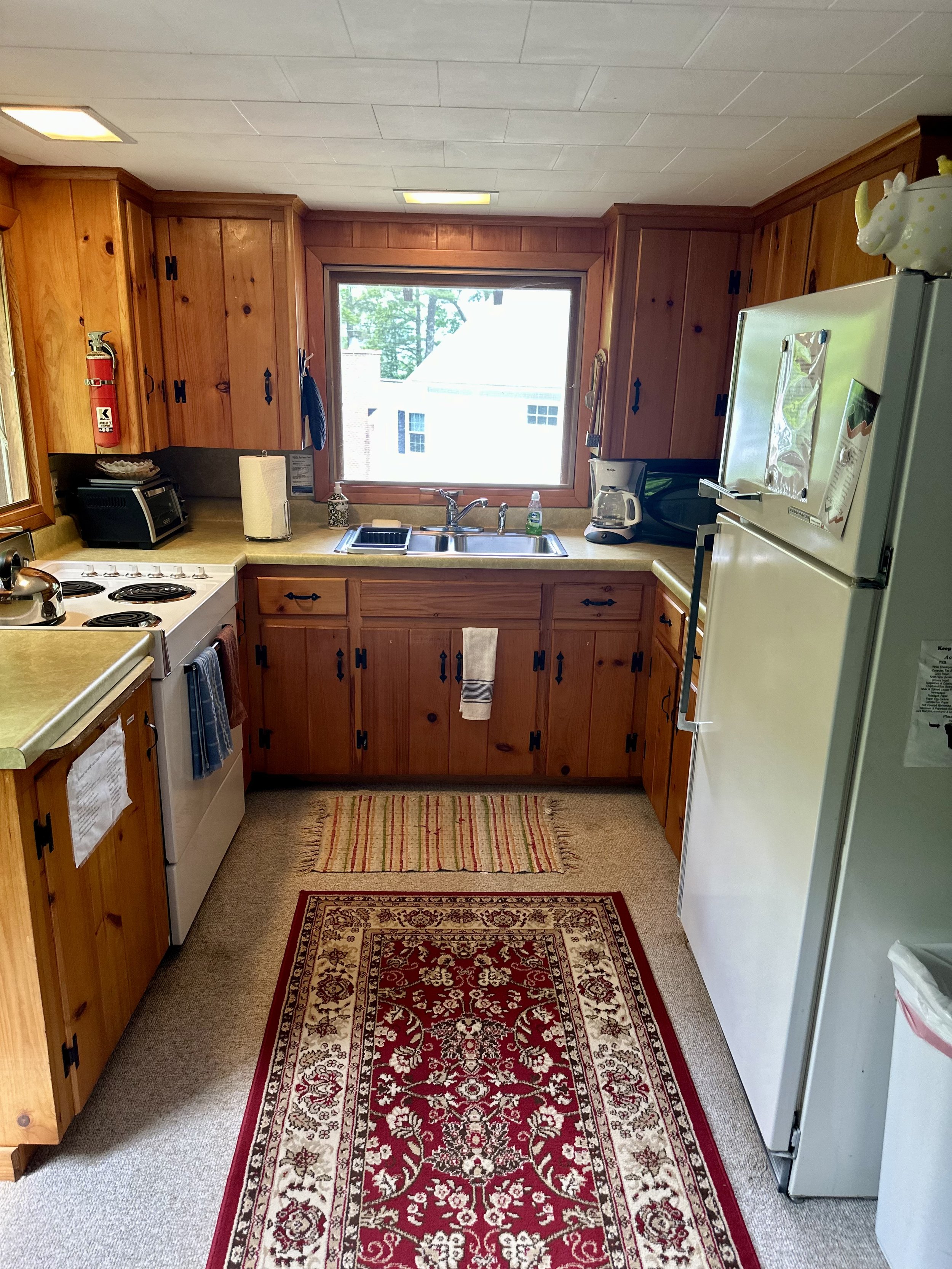 A small kitchen with wooden cabinets, a white refrigerator, a stove, a sink under a window, a small rug with floral patterns, and various kitchen appliances on the countertops.
