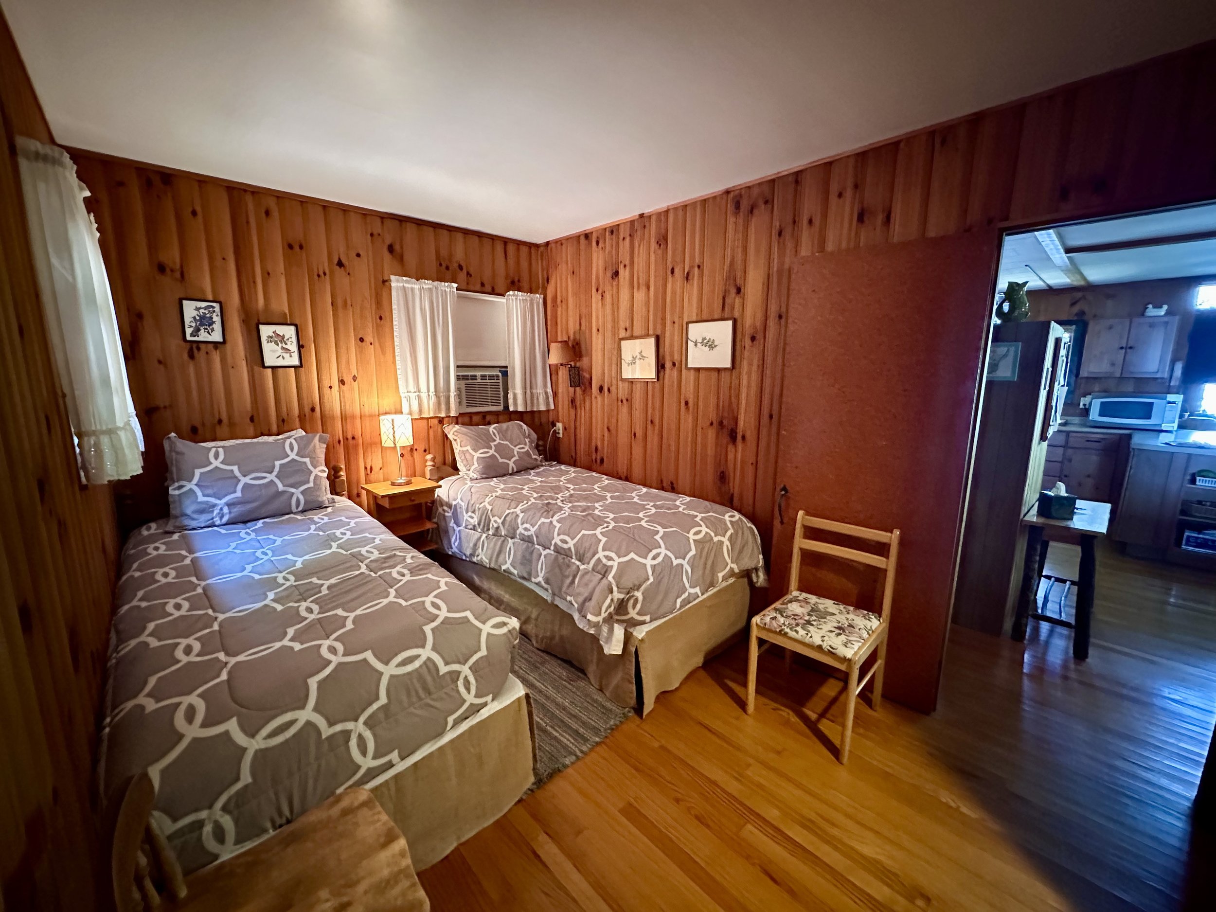 A cozy bedroom with two twin beds featuring patterned beige and white bedding, wooden walls, small picture frames, a window with white curtains, a small wooden nightstand with a lamp, and a wooden chair with a floral cushion. The room opens into a ki