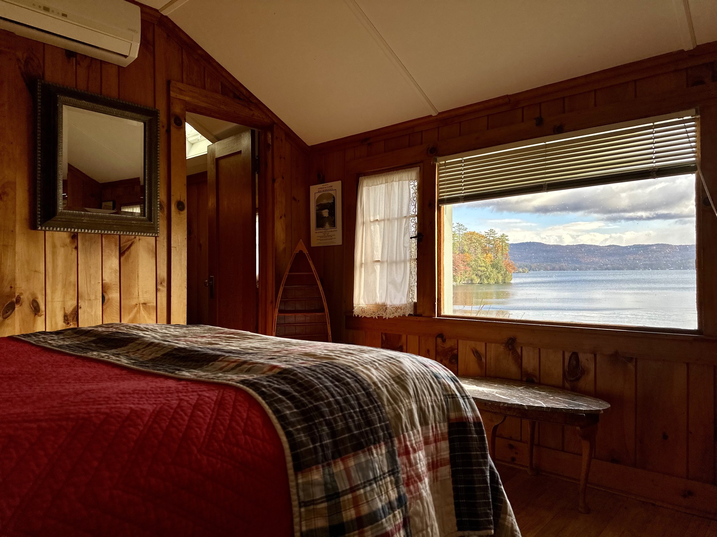 A cozy wooden cabin bedroom with a large window overlooking a lake and trees with autumn foliage, a bed with a plaid quilt, a mirror on the wall, a small bench near the window, and a curtain partially covering a smaller window.