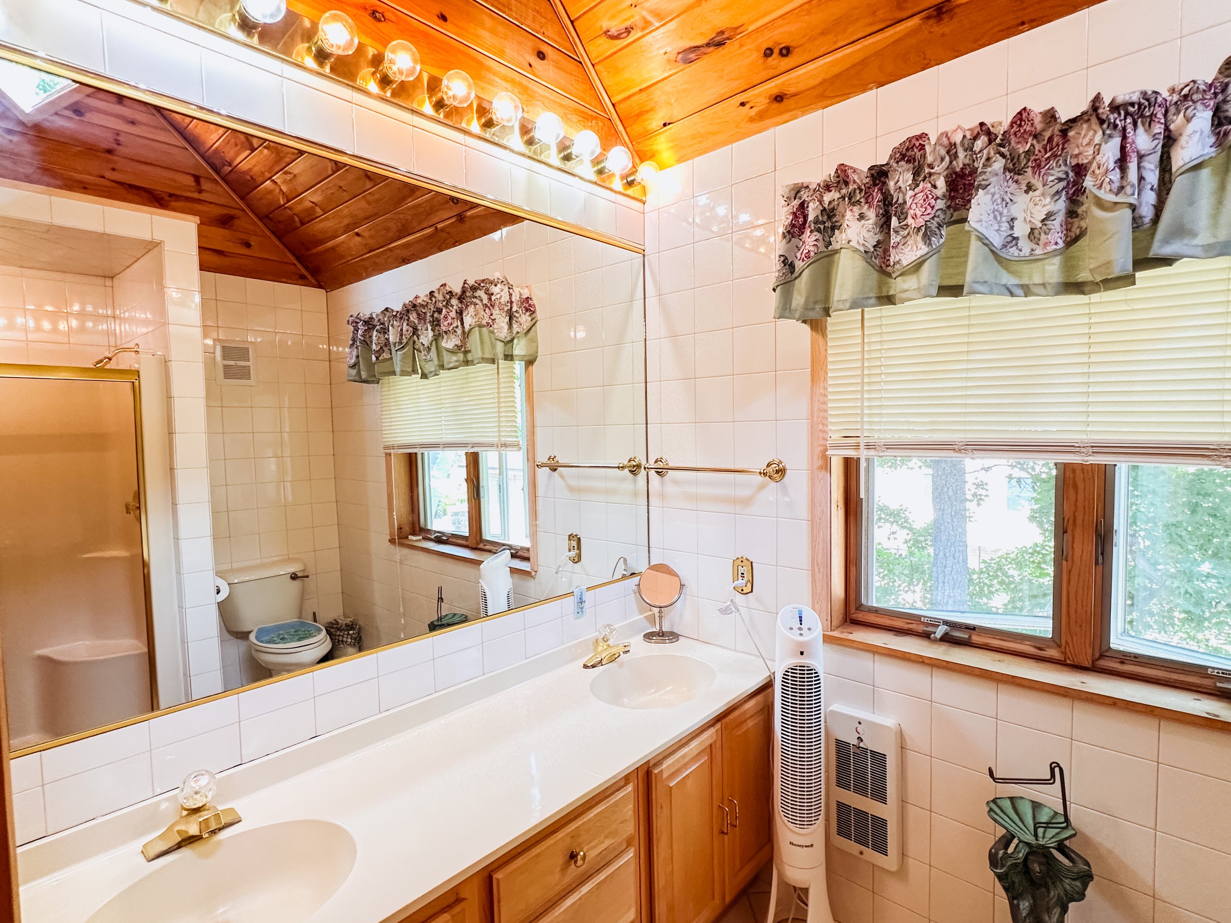 A bathroom with a double sink vanity, a large mirror, and wooden accents. There are two windows with floral valances and blinds, and a ceiling with wood paneling. Various toiletries and a small fan are visible on the sink and countertop.