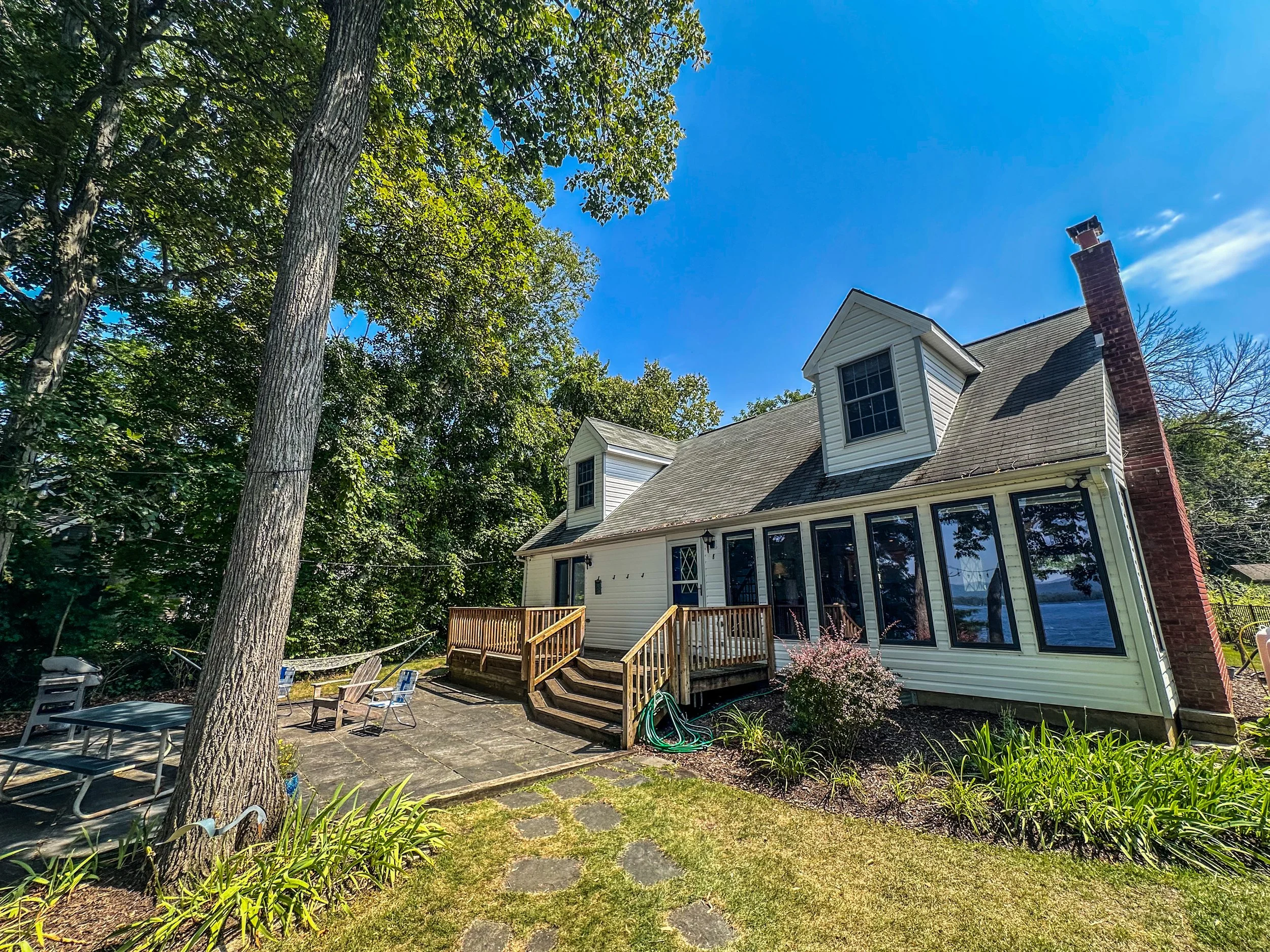 Backyard view of a house with large glass windows, a brick chimney, and a small wooden deck with stairs, surrounded by trees and plants, on a sunny day.