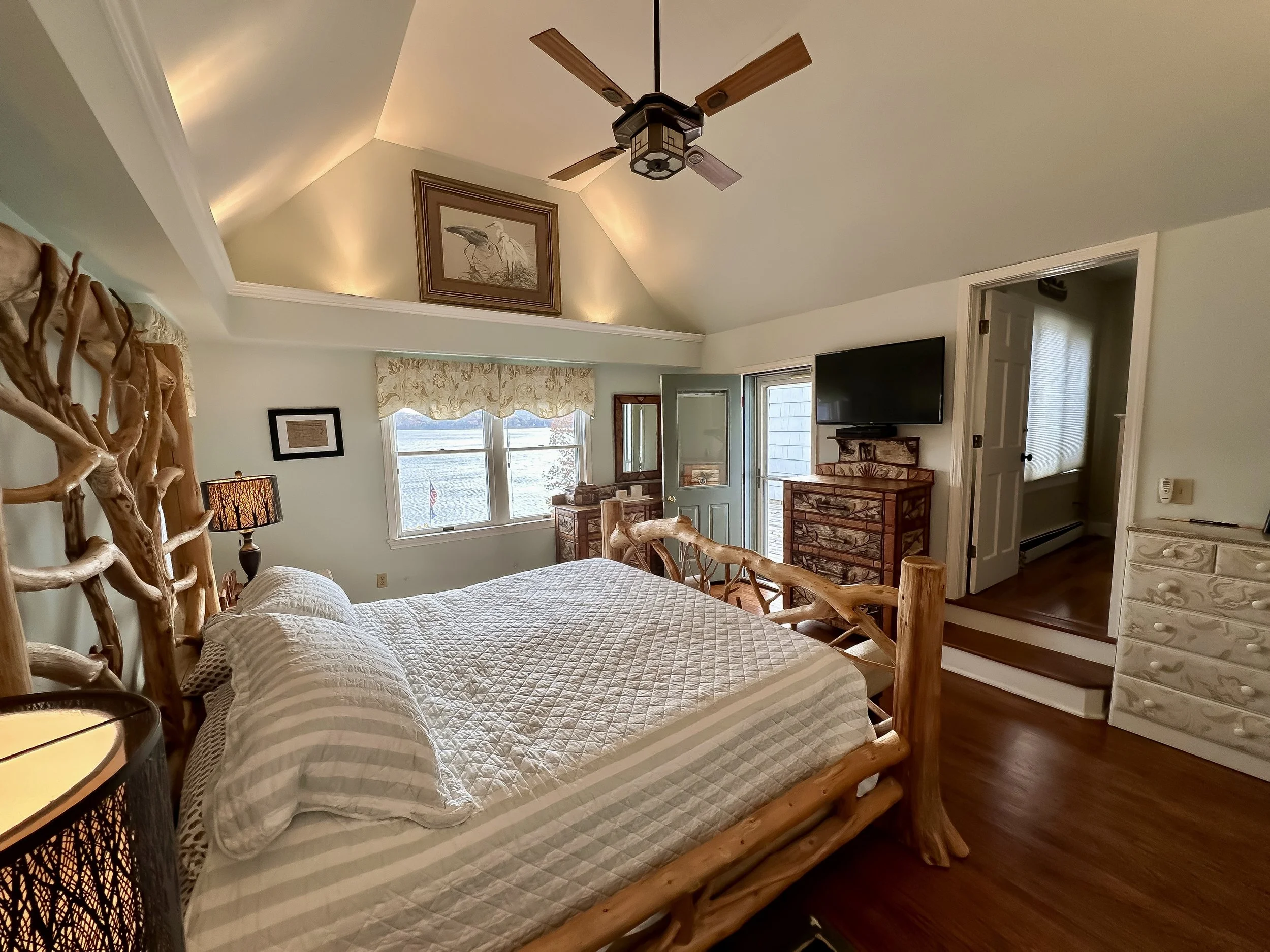 A cozy bedroom with a rustic wooden bed frame, a quilted bedspread, and matching pillows. Features large windows with patterned valances, a ceiling fan, and vintage-style dressers. Contains a wall-mounted flat-screen TV and a decorative framed pictur