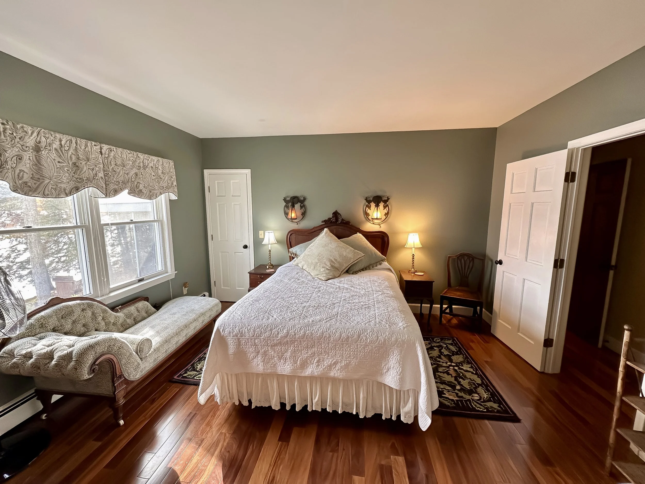 Bedroom with green walls, a large window with a floral valance, vintage chaise lounge, bed with white quilt, two bedside lamps, and two wall sconces above the headboard.