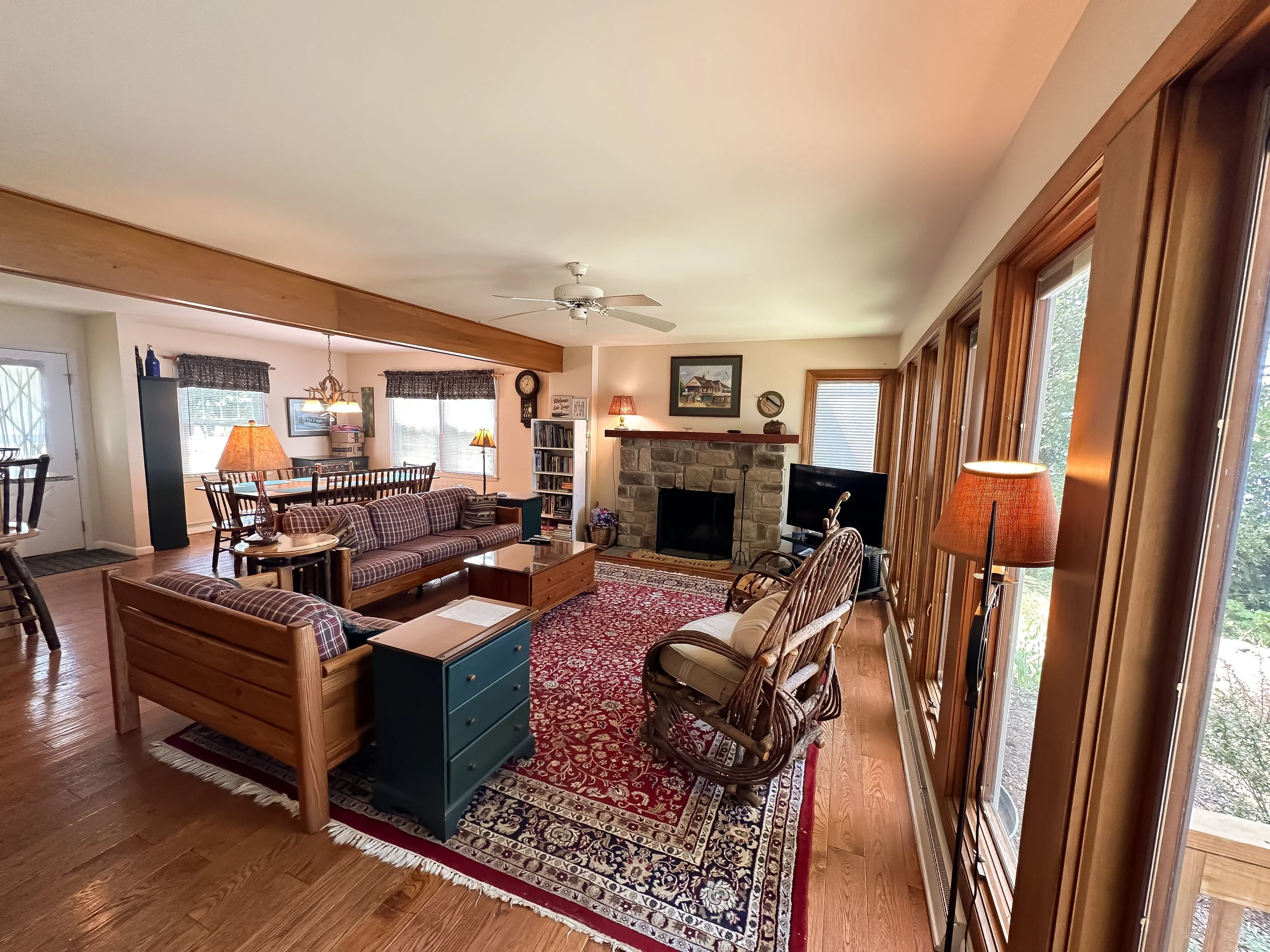 A cozy living room with a brick fireplace, wooden furniture, area rug, large windows with wooden frames, and various seating options, including a checkered sofa and a rocking chair.