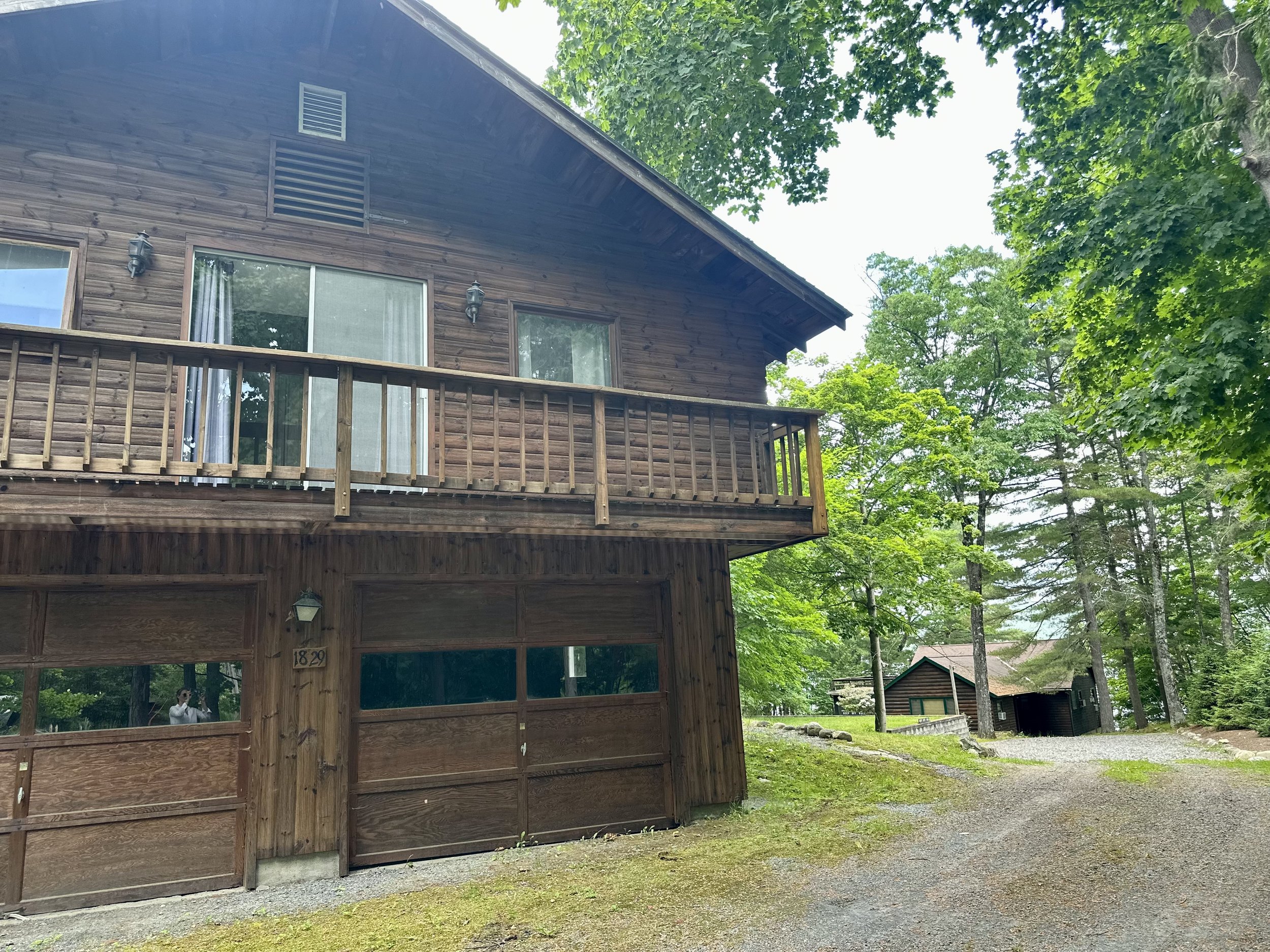A two-story wooden house with a balcony, surrounded by green trees and a gravel driveway.