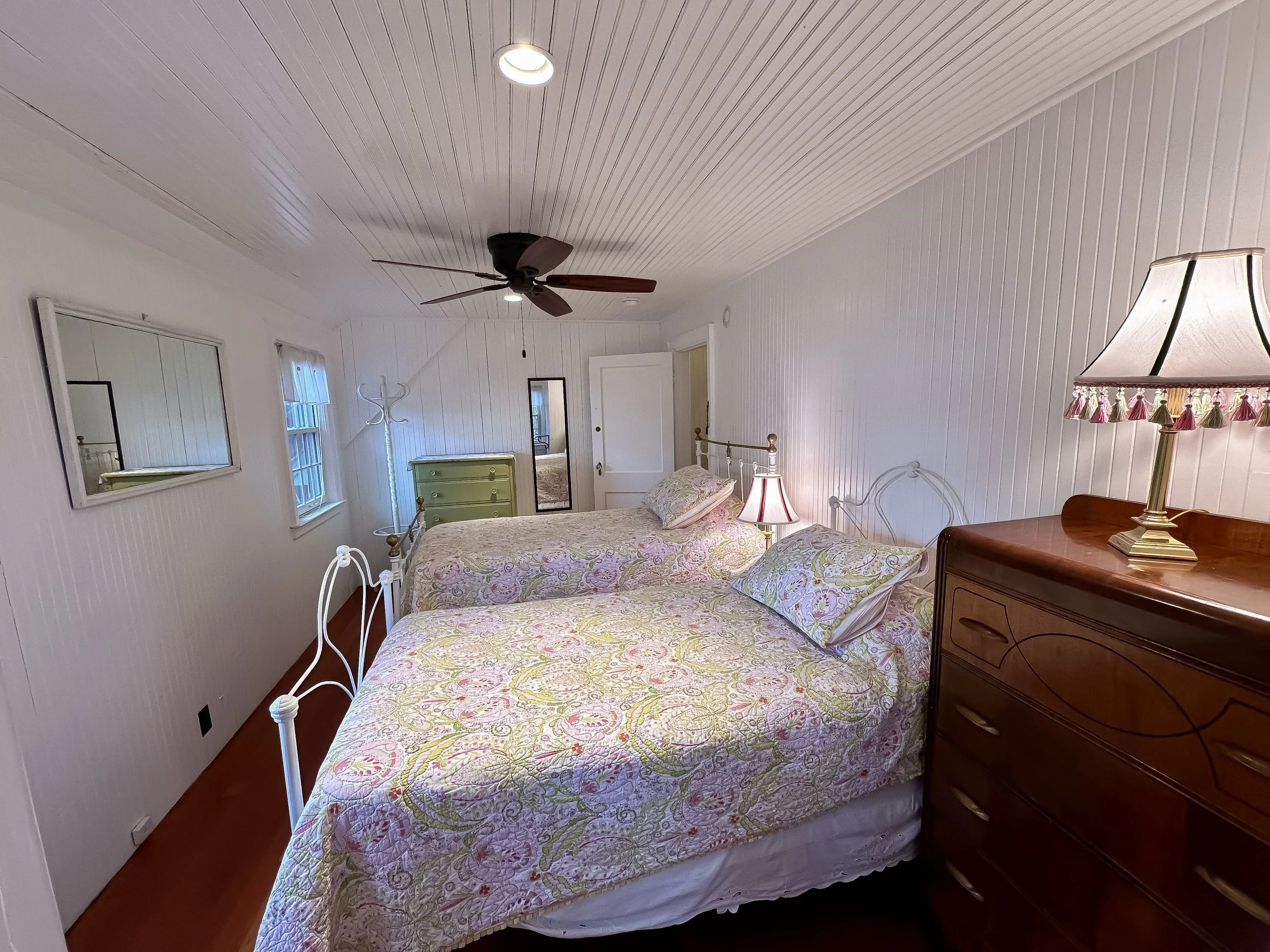 A cozy bedroom with two double beds with floral bedding and pillows, a wooden dresser with a lamp, a mirror, windows, a ceiling fan, and a coat rack.