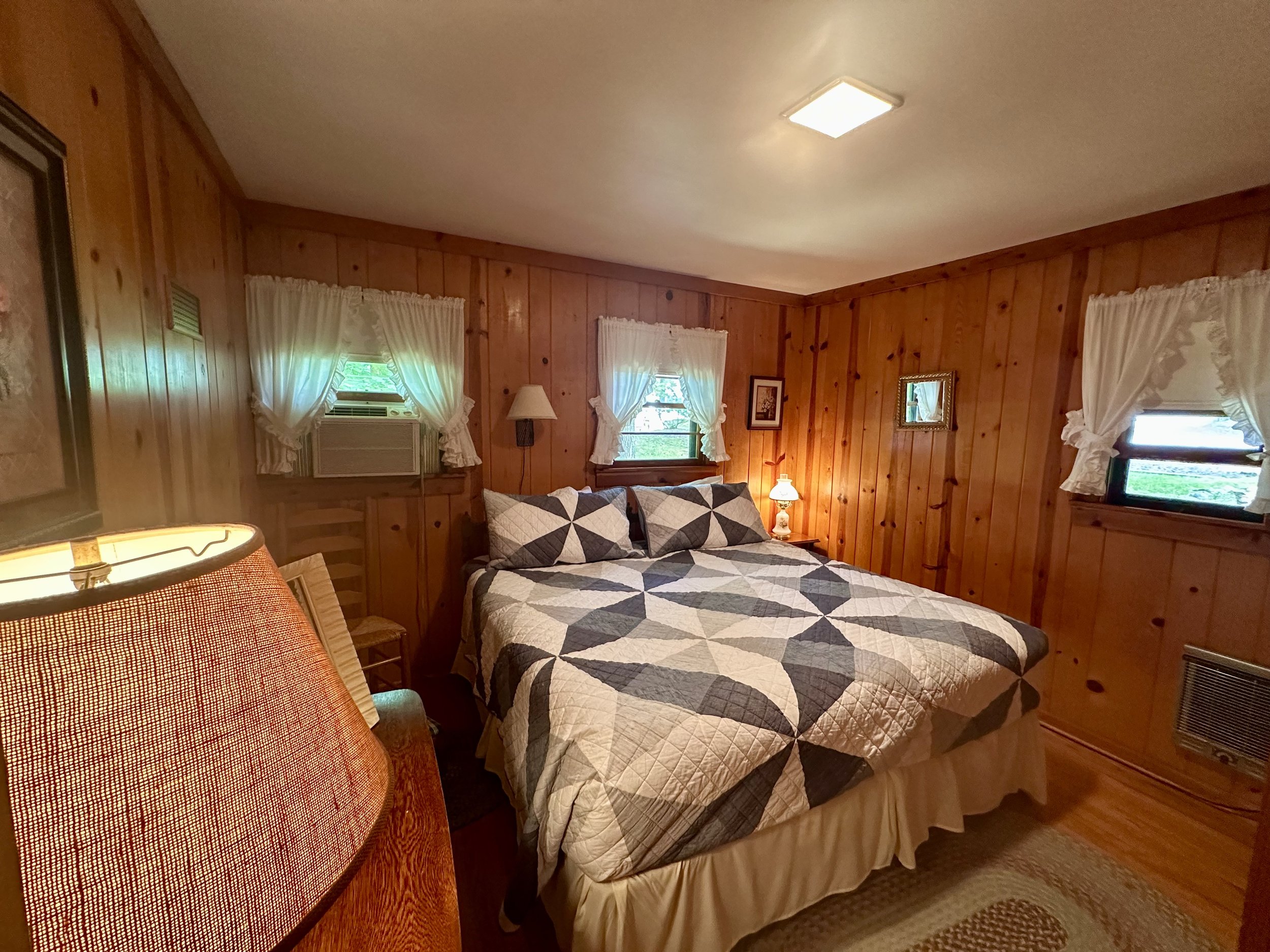 A cozy bedroom with wood-paneled walls, a bed with a geometric quilt, and small windows with white curtains.