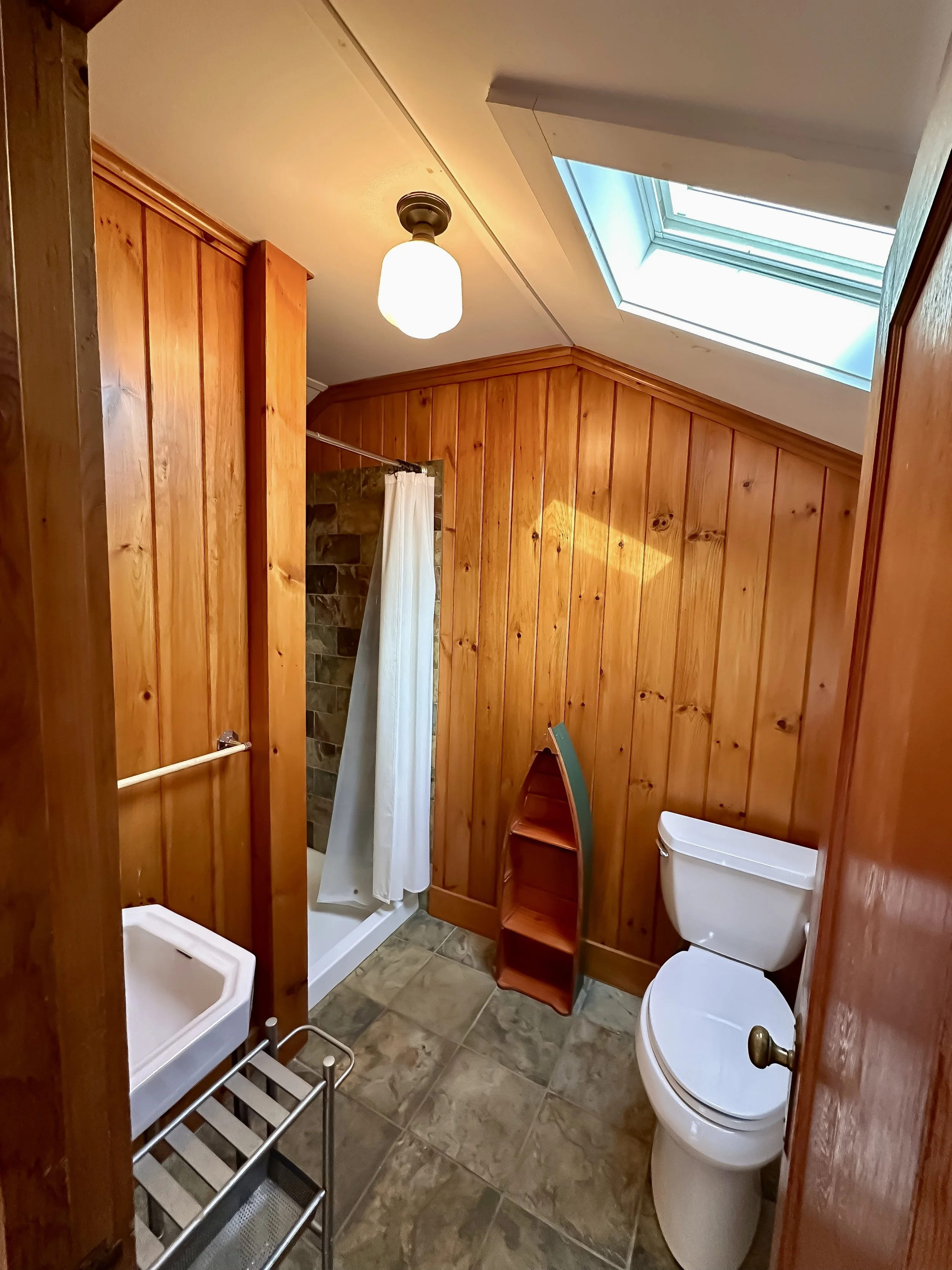 Bathroom with wood-paneled walls, a small corner shower with a white curtain, a white toilet, a small arched shelf, a skylight, and a ceiling light.