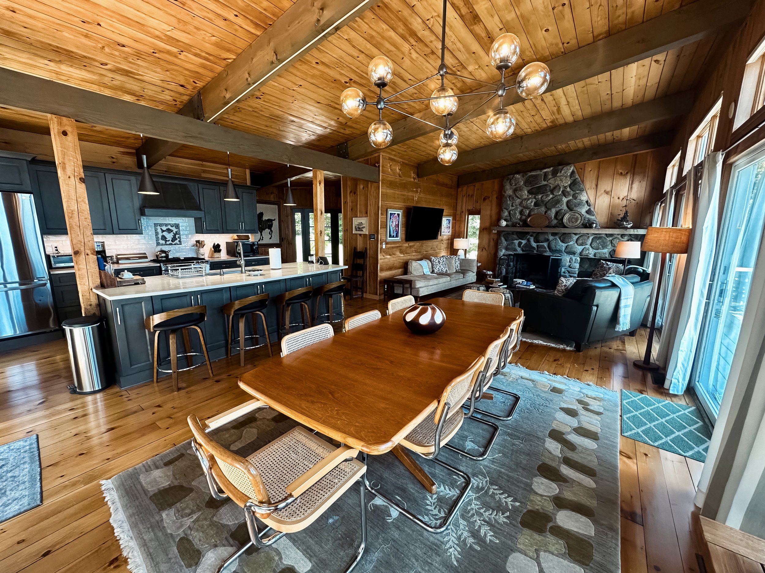 Open-concept living and dining room with wooden ceiling and floors, a long wooden dining table with chairs, a kitchen with a blue island and barstools, a stone fireplace with a flat-screen TV, and a seating area with sofas and lamps near sliding glas