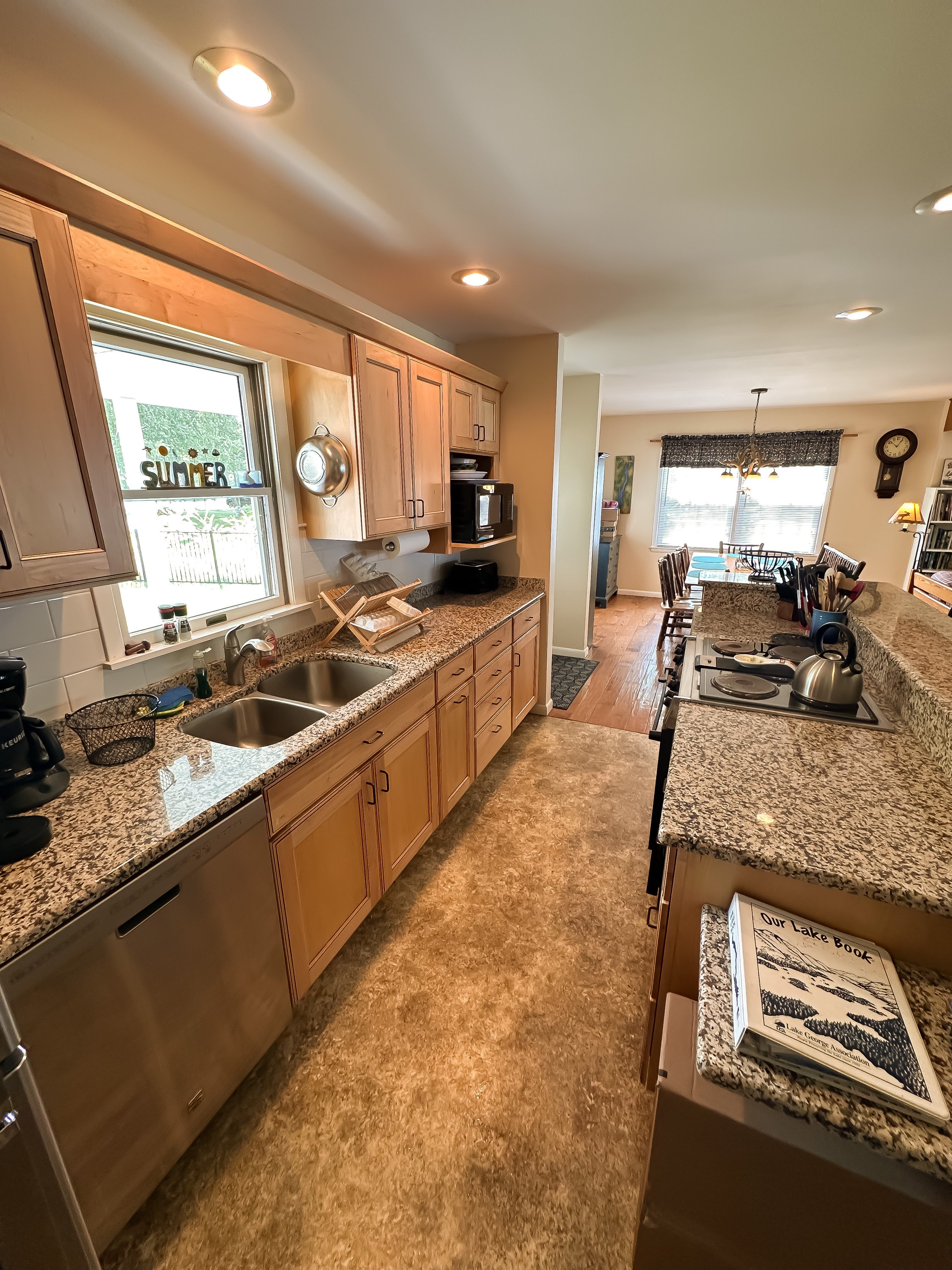 Kitchen with wooden cabinets, granite countertops, stainless steel sink, and a view into the dining area with a table and chairs, large windows, and wall clock.