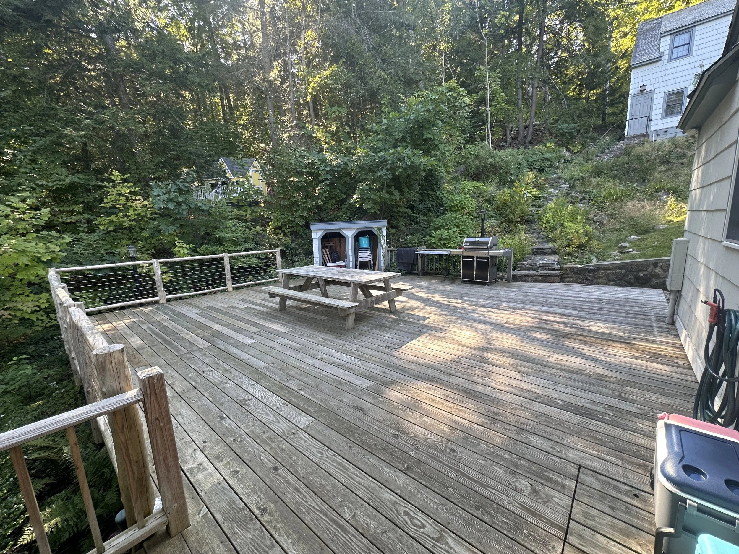 Wooden deck with outdoor grill, picnic table, storage shed, and surrounded by trees and greenery.