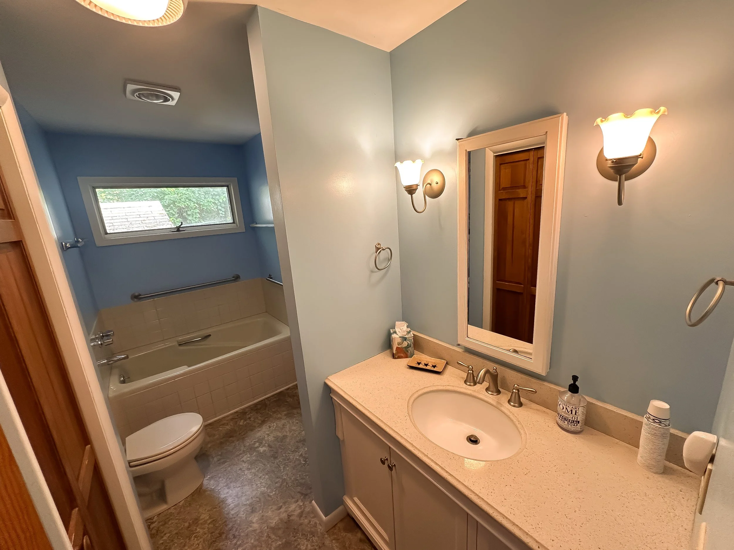 Bathroom with light blue walls, beige vanity with a sink, mirror, wall-mounted lights, and a wood door; bathtub with beige tiles, small window, and a grab bar.